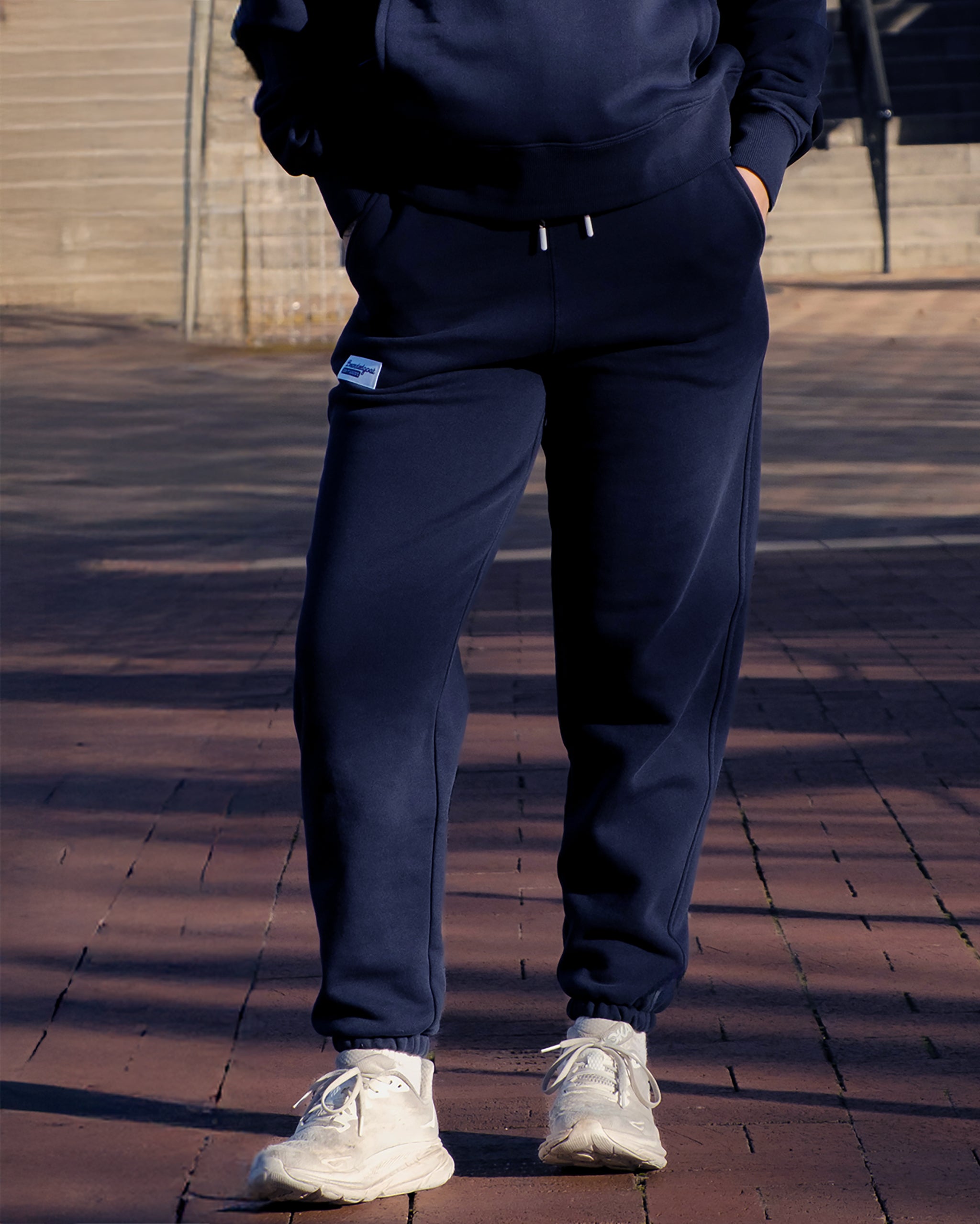 Lower body visible wearing BEARDEDGOAT’s NEW — Overnight™ Classic Jogger in navy blue with matching hoodie, hands in pockets. White sneakers complete the look while standing on brick pavement with steps in the background.