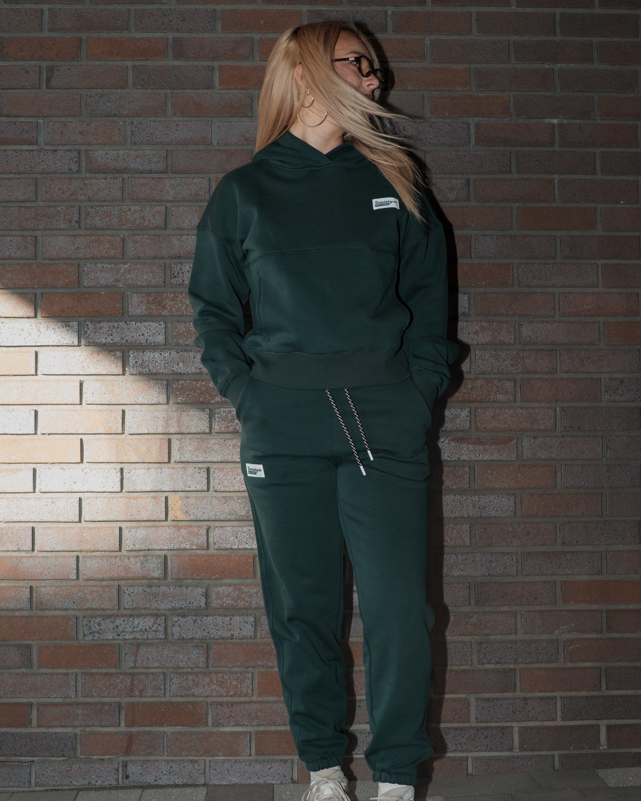 A person with long blonde hair stands against a brick wall, wearing BEARDEDGOAT's NEW — Overnight™ Classic Jogger made from heavyweight cotton fleece. Hands in pockets, they look to the side as their hair blows in the wind.