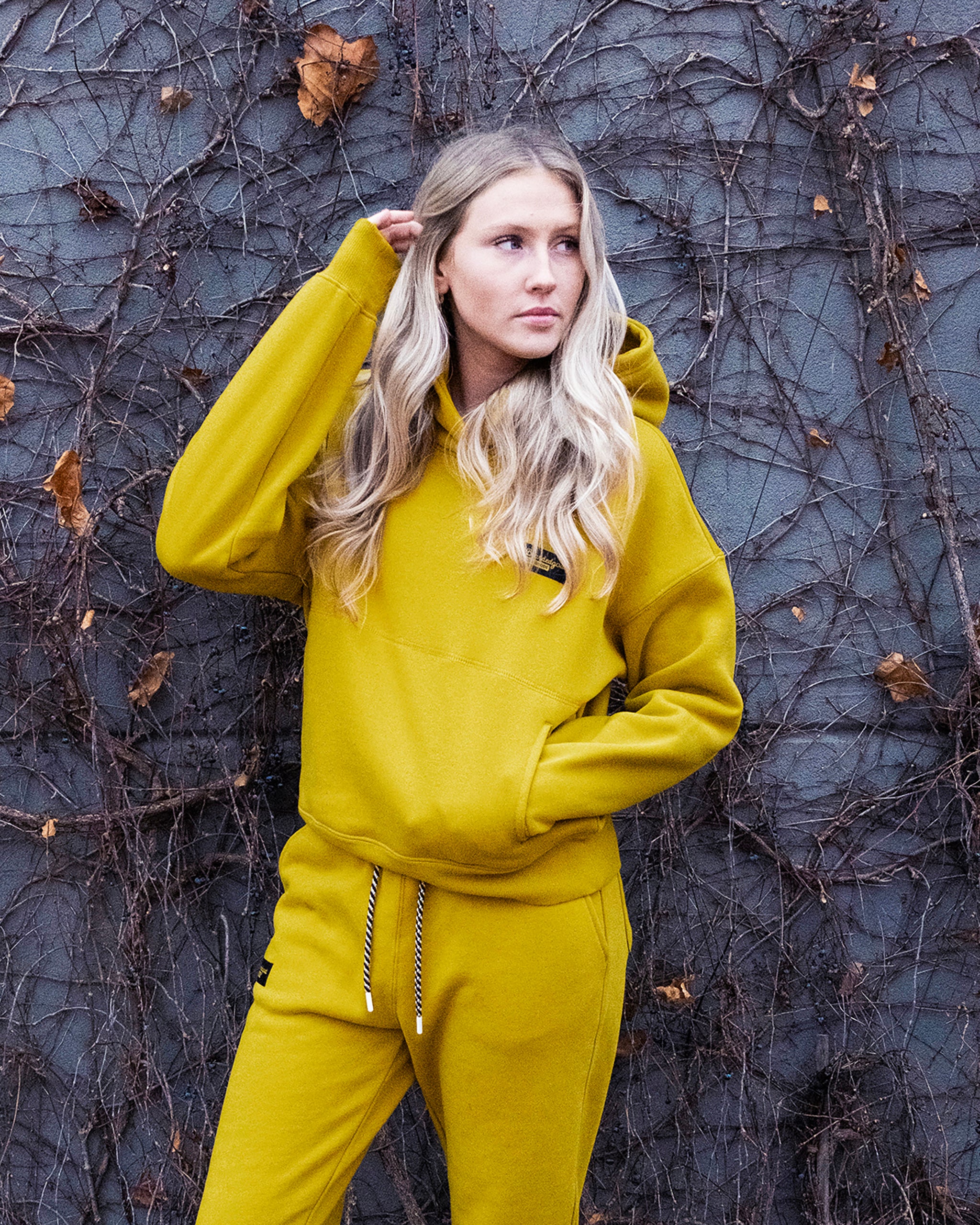 A woman with long blonde hair wears the beardedgoat Women's Overnight™ Hoodie and matching pants, made from heavyweight cotton fleece, standing before a gray vine-covered wall and looking to the side with one hand near her head.
