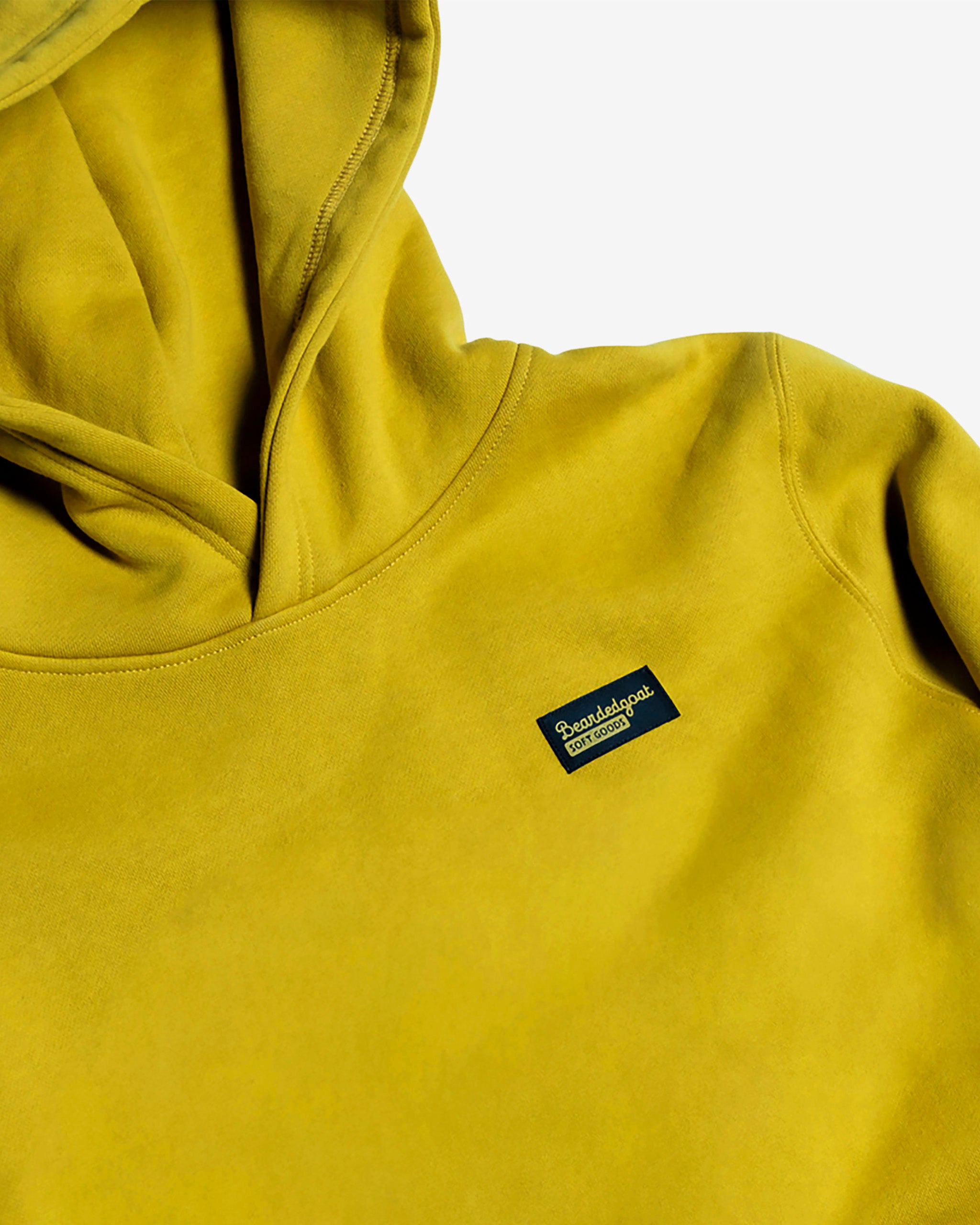 A mustard yellow Women's Overnight™ Hoodie by beardedgoat, crafted from heavyweight cotton fleece with a small rectangular black and yellow Paradigm logo patch on the chest, shown against a plain white background.