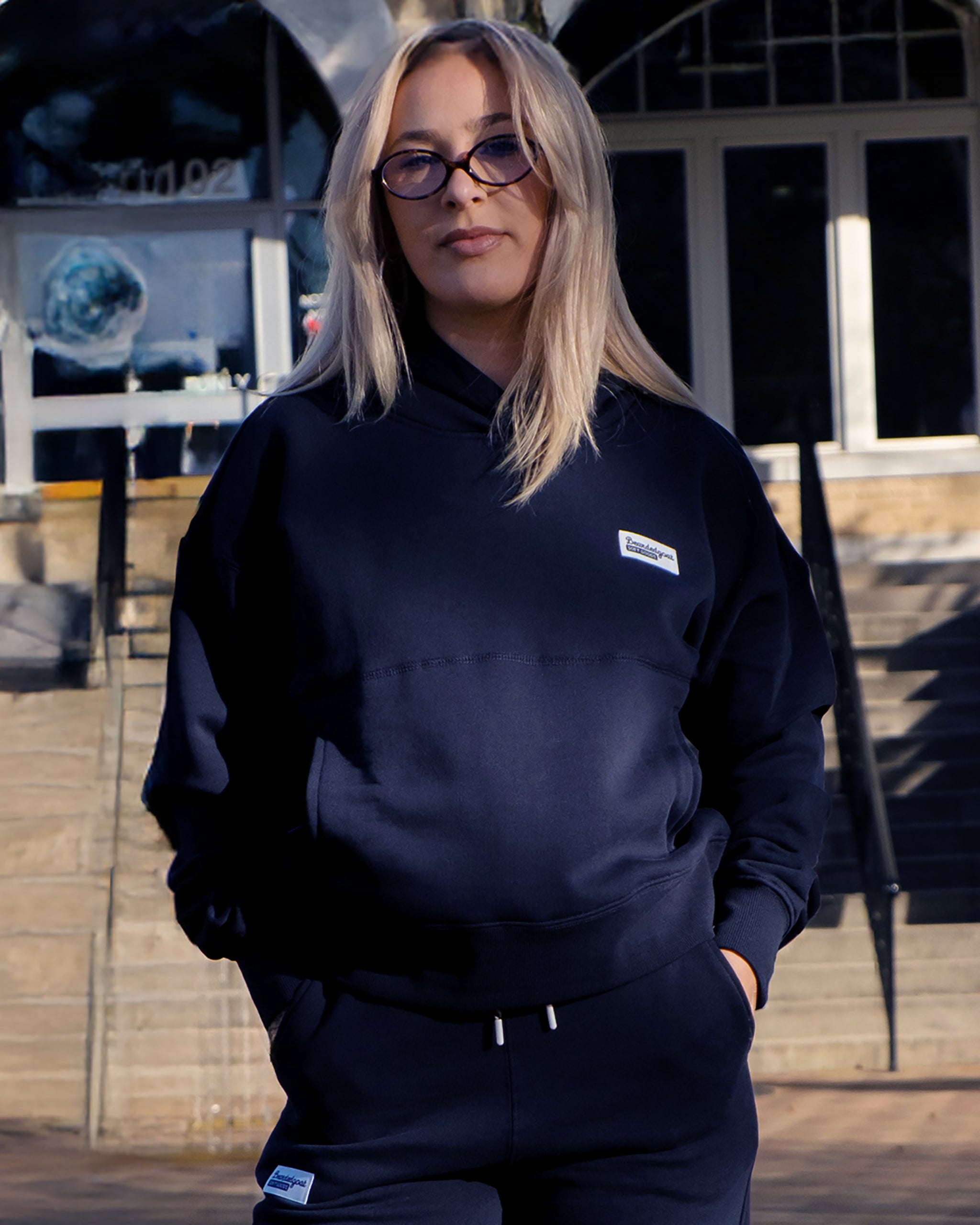 A woman with long blonde hair and glasses wears the beardedgoat Women's Overnight™ Hoodie, crafted from premium cotton fleece, as she stands outside in front of steps and glass doors.