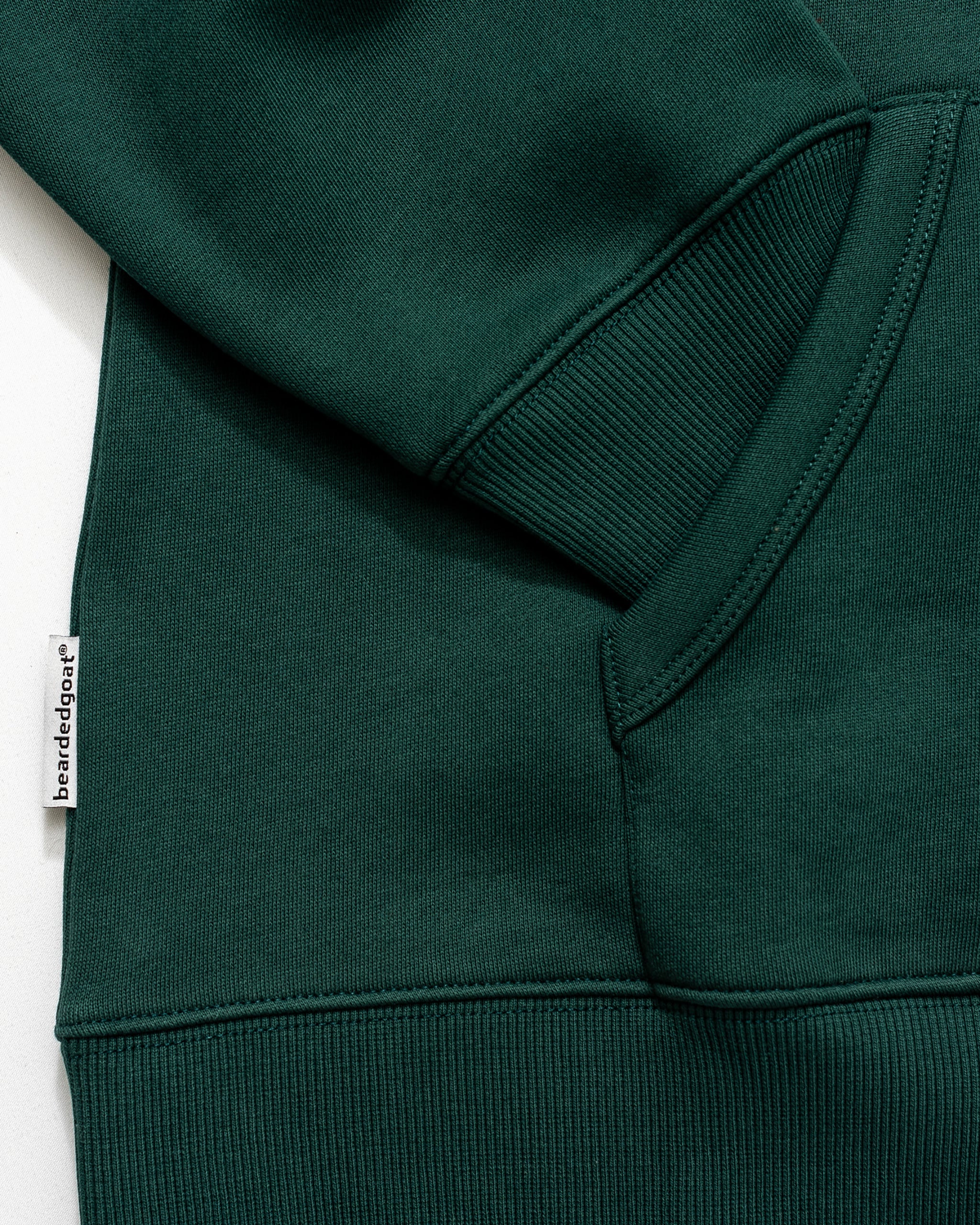 Close-up of the BEARDEDGOAT NEW — Overnight™ Half Zip, a dark green heavyweight fleece sweatshirt featuring a ribbed cuff, kangaroo pocket, and a small white side tag reading "brandedgar.