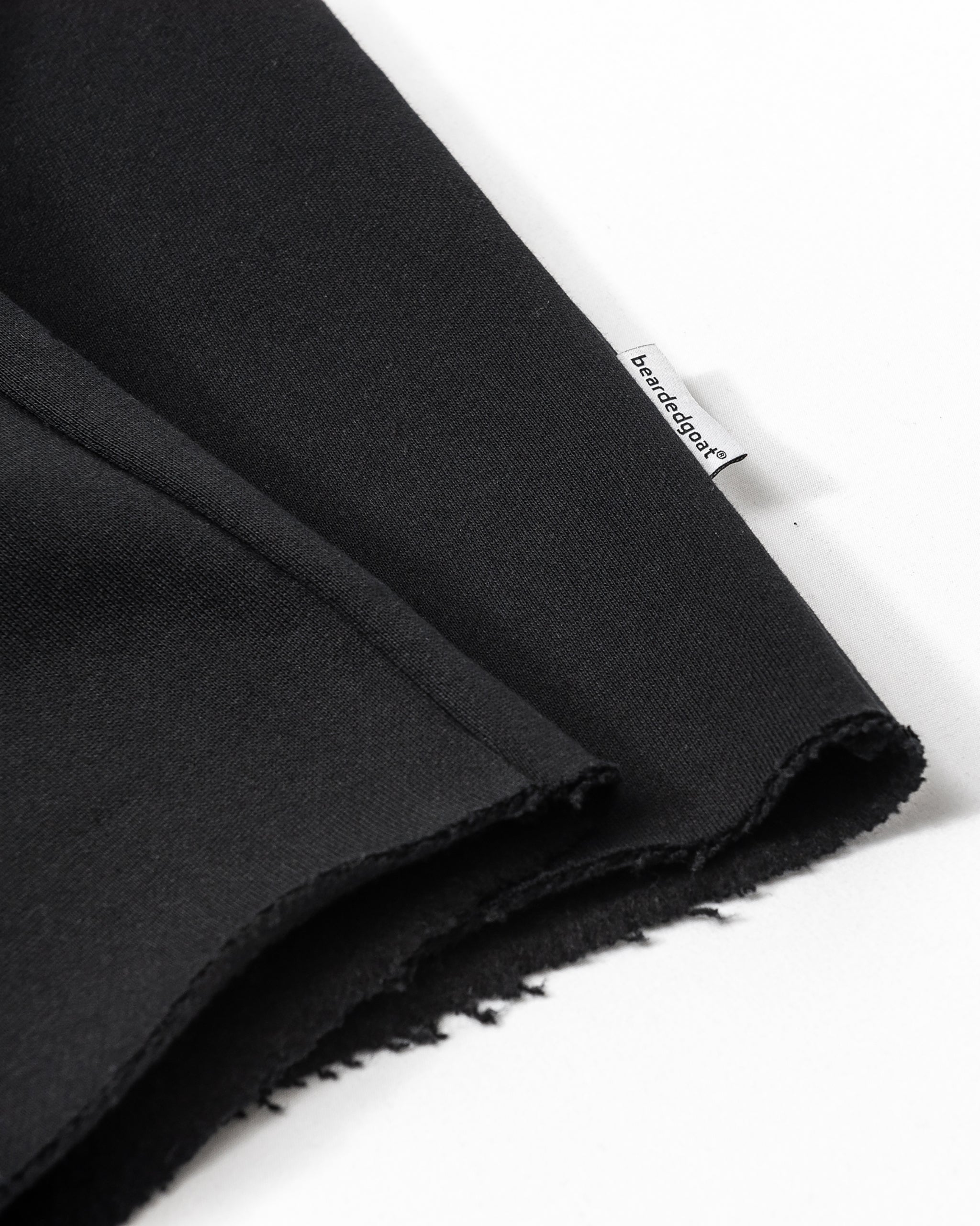 A close-up of black heavyweight cotton fleece with raw edges, featuring a small white beardedgoat tag sewn into the seam—ideal for the Women’s Overnight Sweatpant—displayed on a white surface.