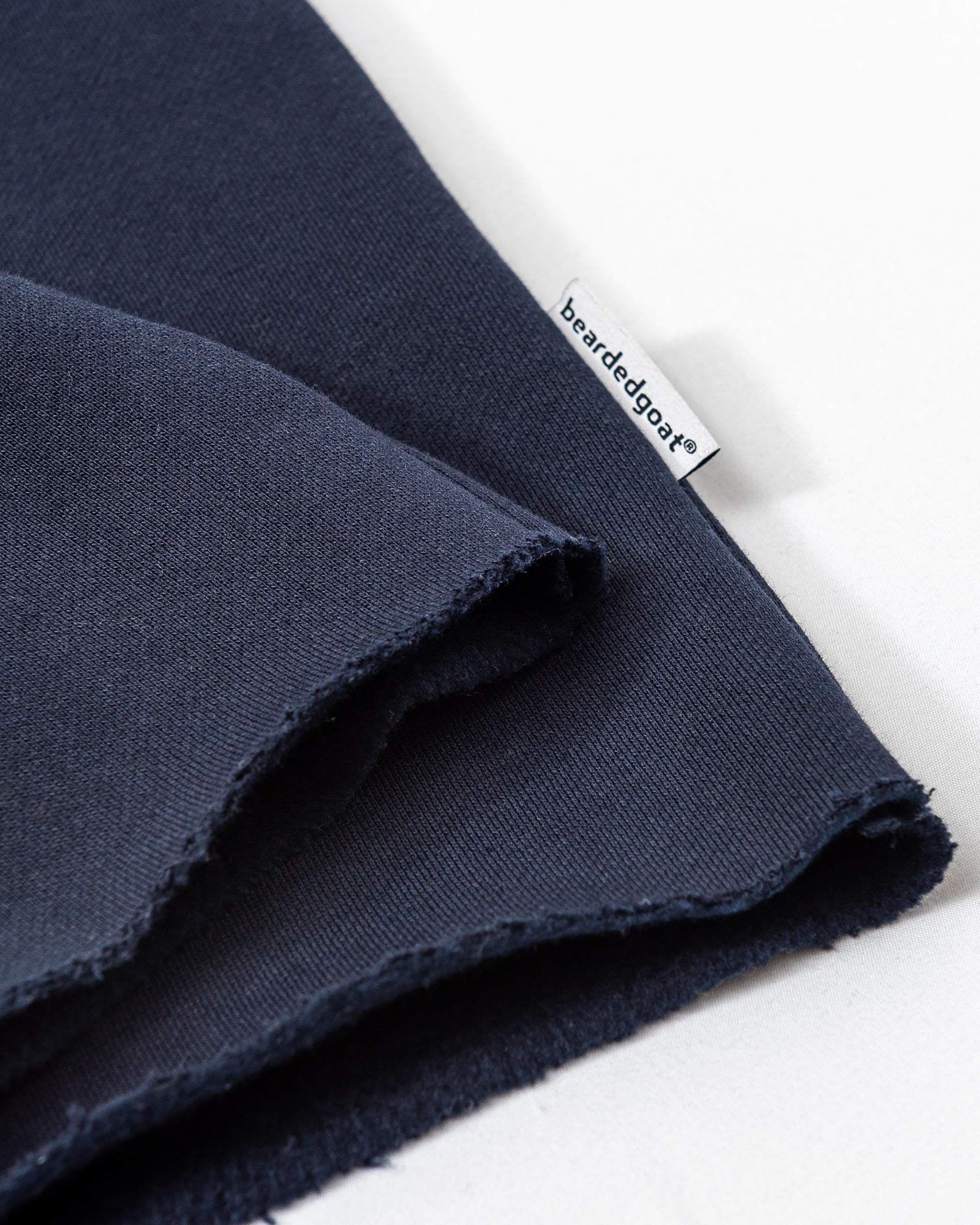 A close-up of heavyweight dark blue cotton fleece with raw edges, featuring a small white beardedgoat tag. The fabric, perfect for the Women’s Overnight Sweatpant, is laid on a white surface.