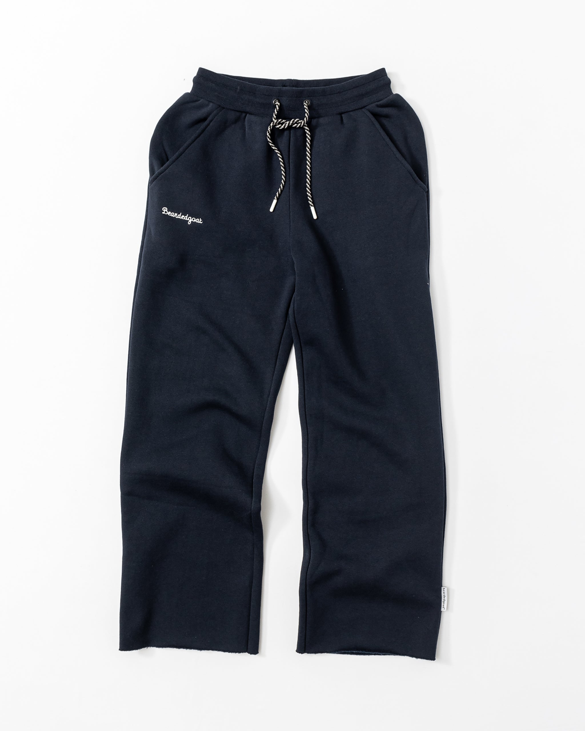 The beardedgoat Women’s Overnight Sweatpant in navy blue is made from heavyweight cotton fleece, with a white drawstring, side pockets, and small white embroidered text on the left thigh, shown laid flat on a white background.