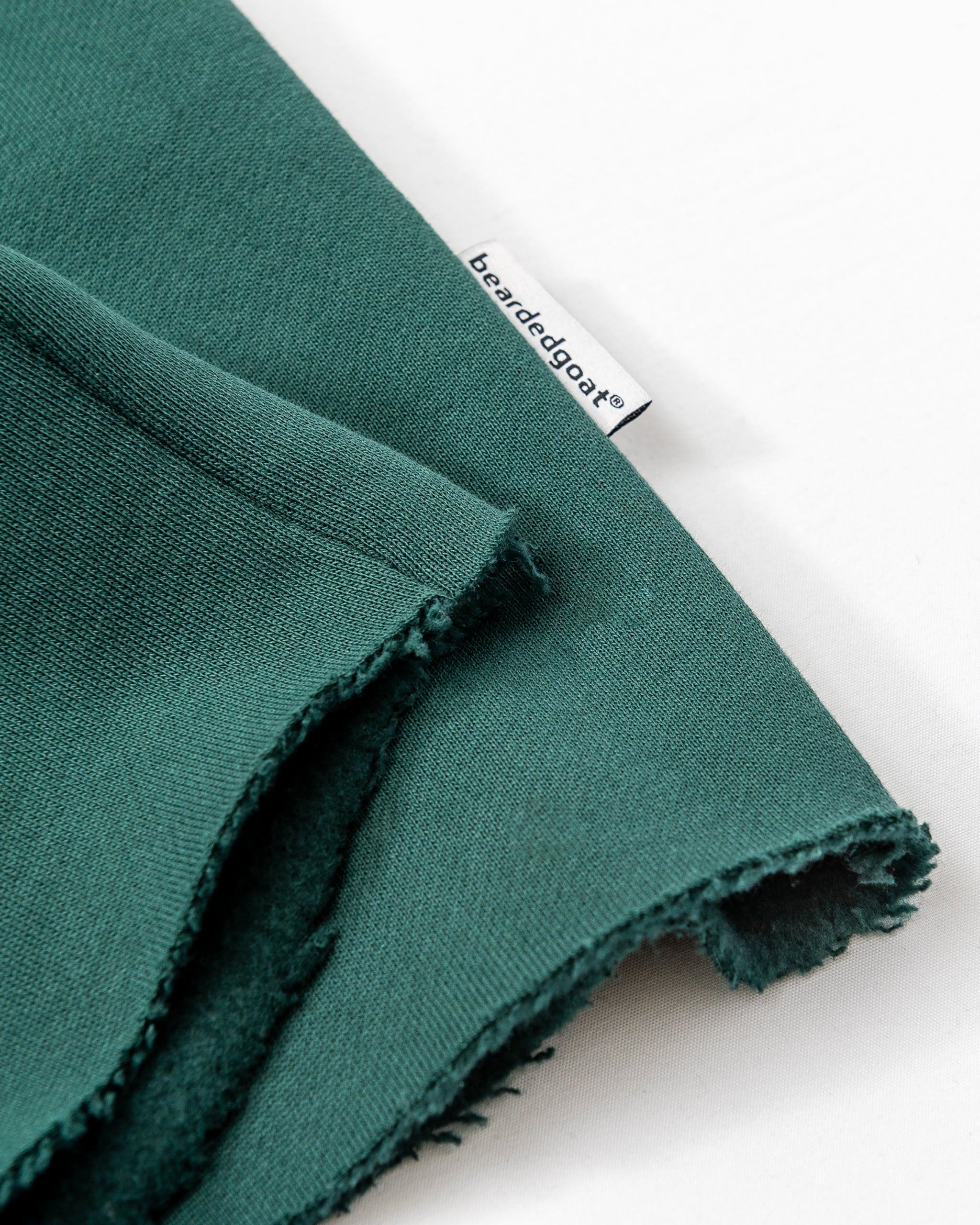 Close-up of heavyweight cotton fleece with raw edges and a sewn-in white beardedgoat tag. The fabric, shown flat on a white surface, is used for the Women’s Overnight Sweatpant by beardedgoat—ideal for comfortable loungewear styles.