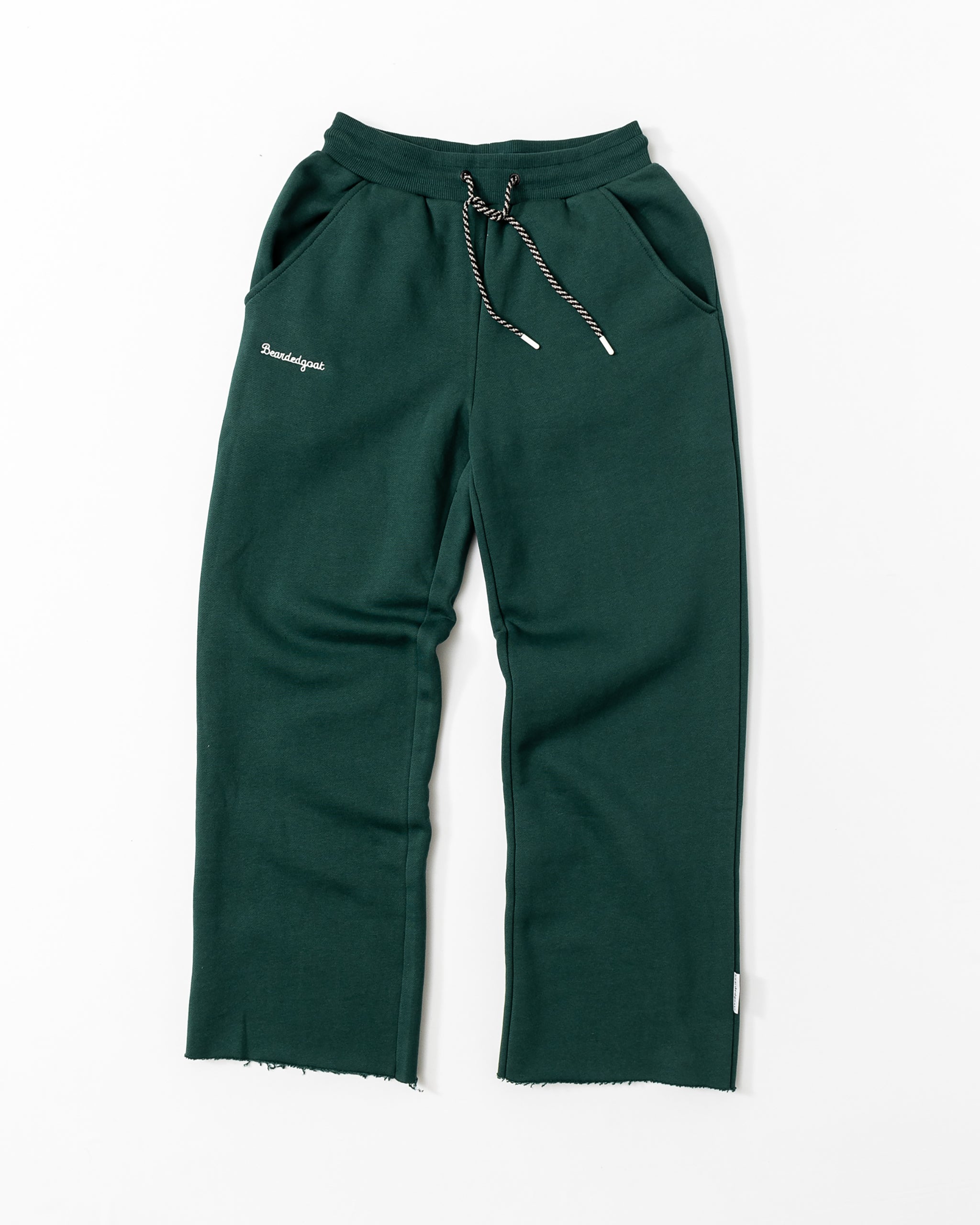 The beardedgoat Women’s Overnight Sweatpant in dark green is made from heavyweight cotton fleece, with a drawstring waist, side pockets, raw edge hems, and small white Paradigm embroidery on the left hip. Shown laid flat on a white background.