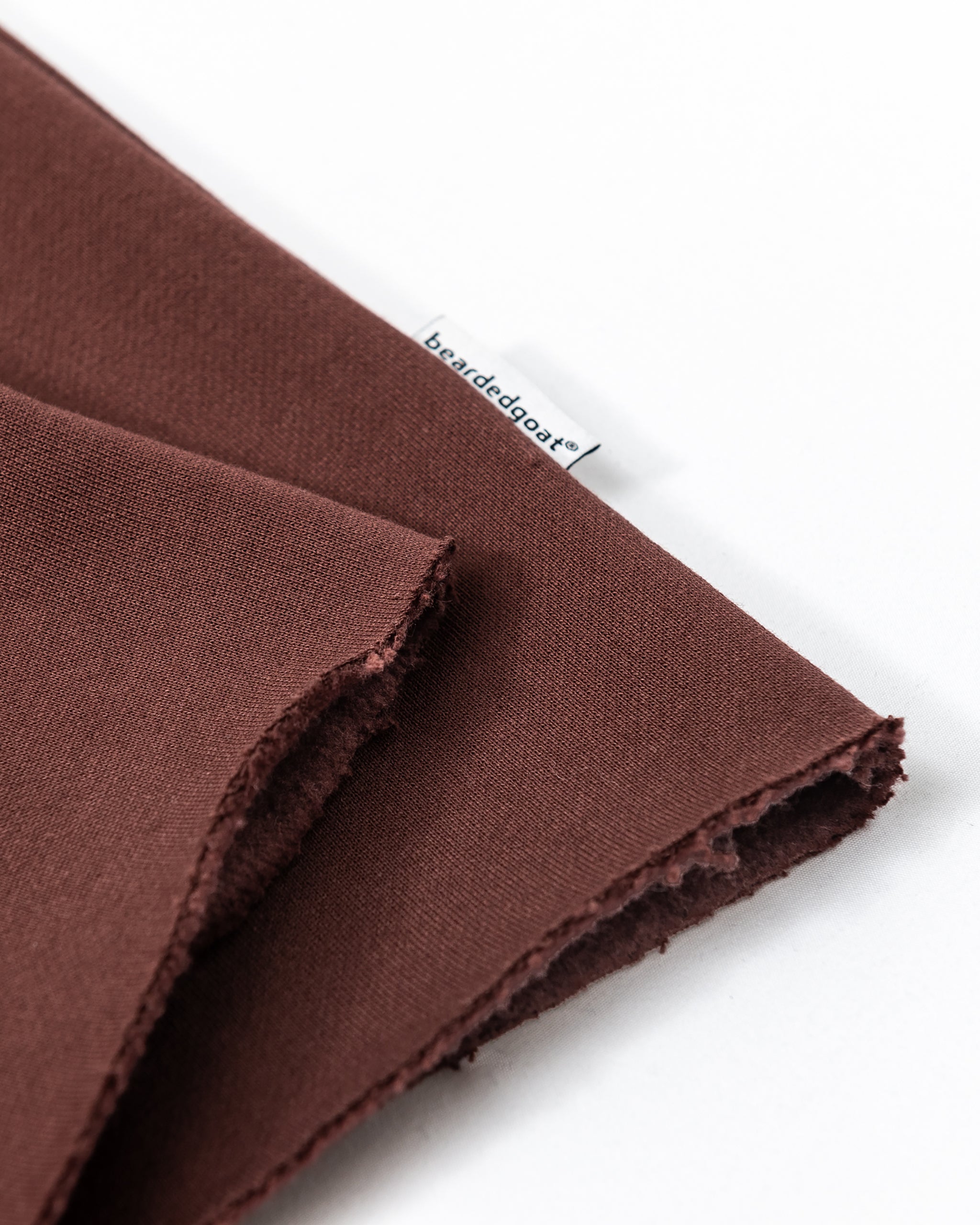Two pieces of brown heavyweight cotton fleece, including the Women’s Overnight Sweatpant by beardedgoat, overlap on a white surface with slightly frayed edges. A small white tag with blue text shows the beardedgoat brand.