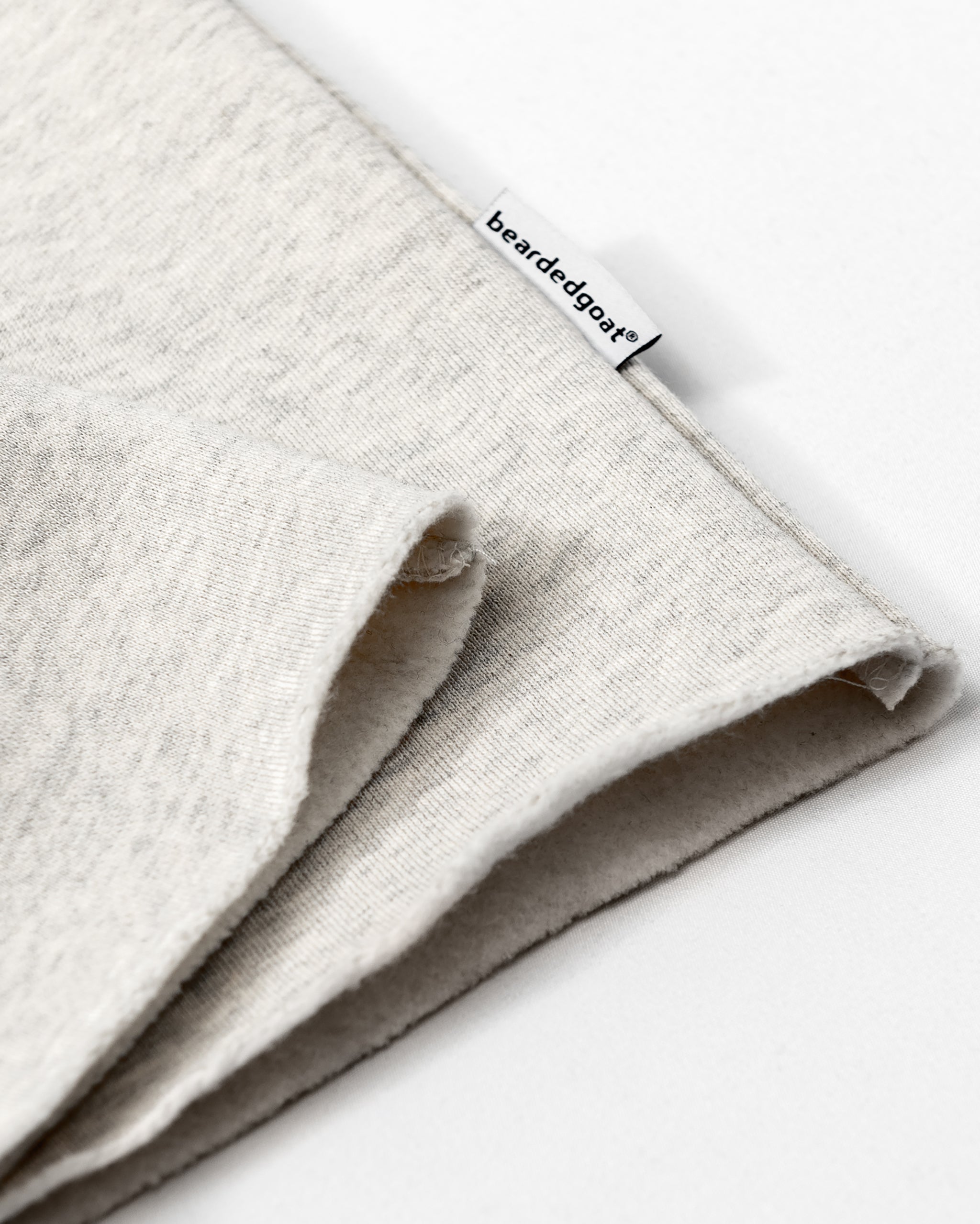 Close-up of a soft, light gray heavyweight cotton fleece from the beardedgoat Women’s Overnight Sweatpant, folded with a small white beardedgoat® tag on the seam, set against a white background.