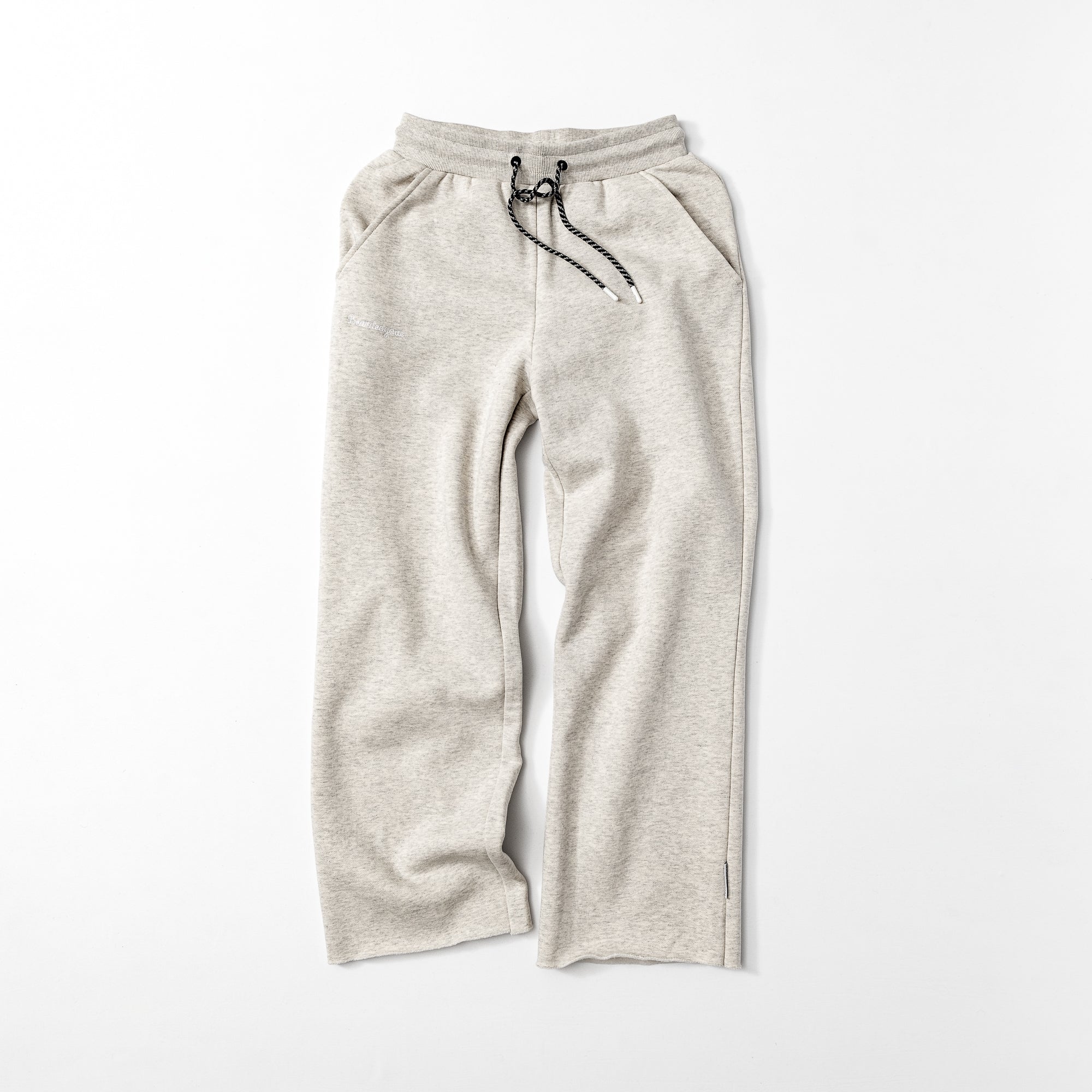 The beardedgoat Women’s Overnight Sweatpant in light gray is made from heavyweight cotton fleece, featuring an elastic waistband with black drawstring, side pockets, and straight legs. Displayed laid flat on a white background.