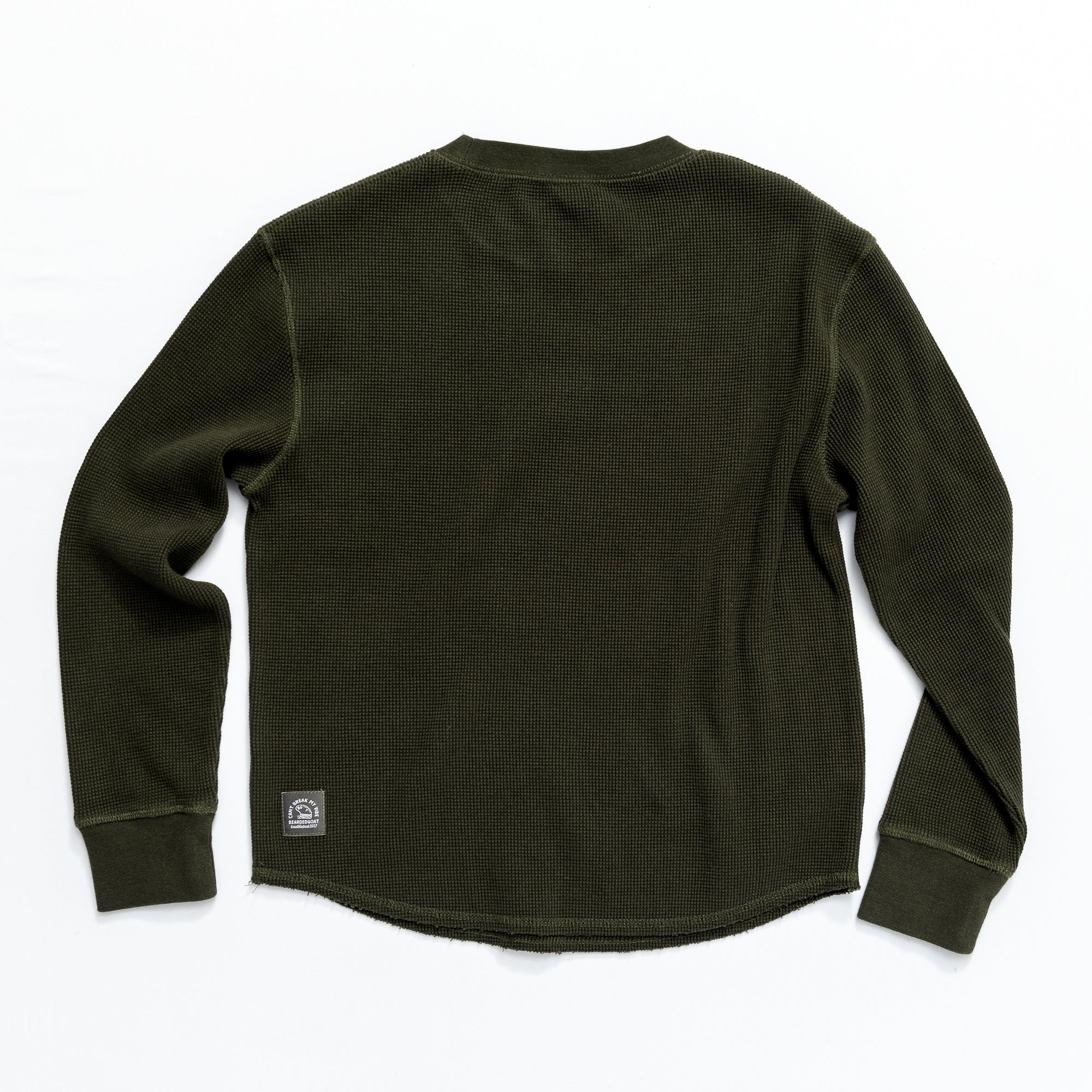 The Womens Wakeside Waffle by BEARDEDGOAT is a dark green cotton long-sleeve, shown laid flat from the back with ribbed cuffs and a small rectangular patch near the hem for a vintage-inspired look.