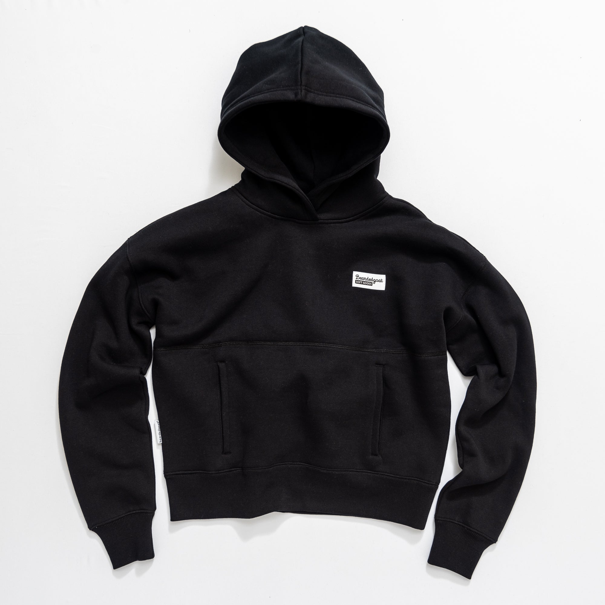 The beardedgoat Women's Overnight Hoodie is a heavyweight cotton fleece in plain black, with a front pocket and a small white rectangular chest patch, shown laid flat on a white background.