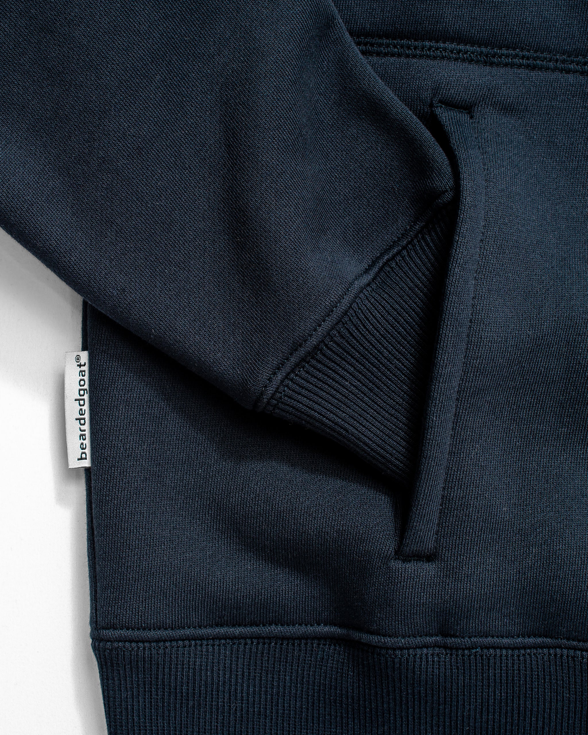 Close-up of the NEW — VOID™ Half Zip sweatshirt by BEARDEDGOAT in dark blue premium cotton fleece, featuring a sleeve, ribbed cuff, kangaroo pocket, and a small white fabric tag with "barelédoga" sewn into the side seam.