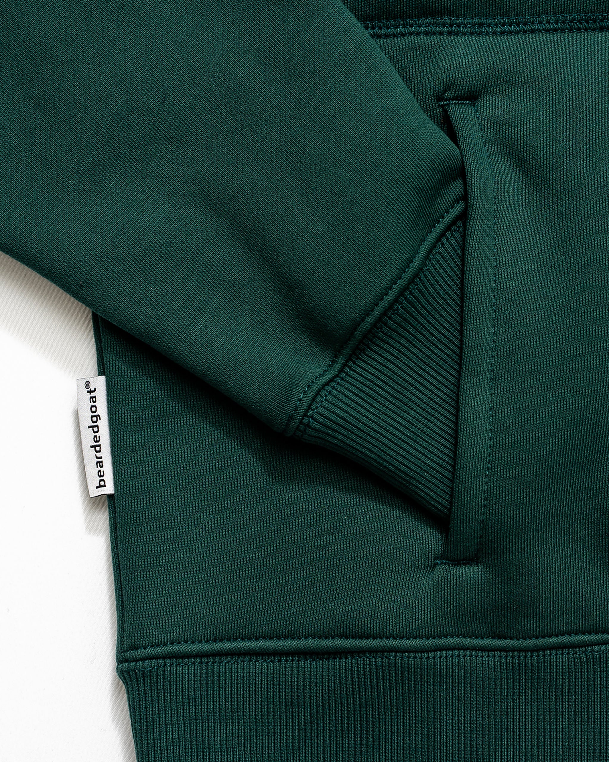 Close-up of a dark green heavyweight NEW — VOID™ Half Zip sweatshirt by BEARDEDGOAT, featuring a ribbed cuff, kangaroo pocket, and a white tag with BEARDEDGOAT branding sewn into the side seam.