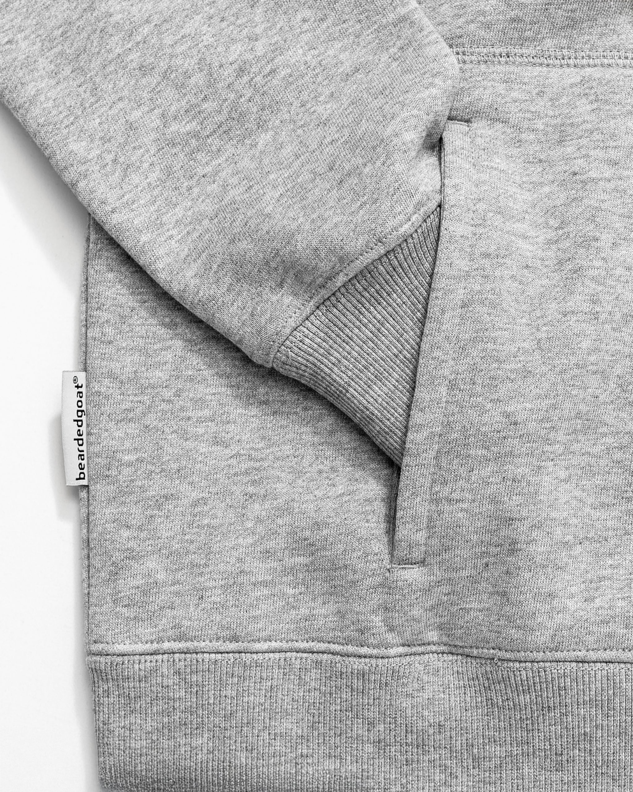 A close-up of the BEARDEDGOAT NEW — VOID™ Half Zip in light gray cotton fleece highlights the cuffed sleeve, front pocket, and a small "blenderdeger" fabric tag sewn into the side seam.