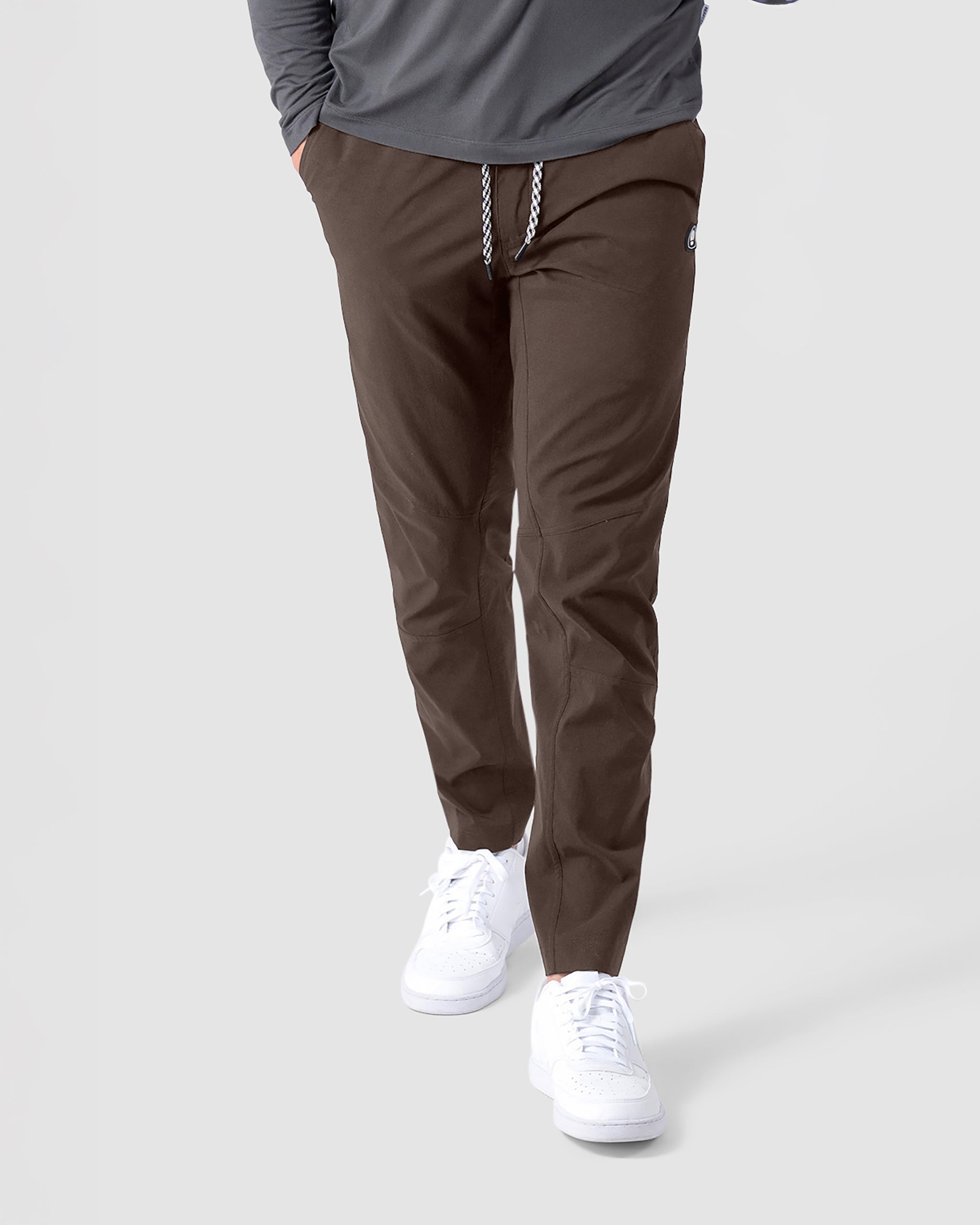 A person stands against a plain light background, wearing BEARDEDGOAT's NEW—ROVER™ Pant in brown with a braided drawstring, partnered with white sneakers and a gray long-sleeve shirt.