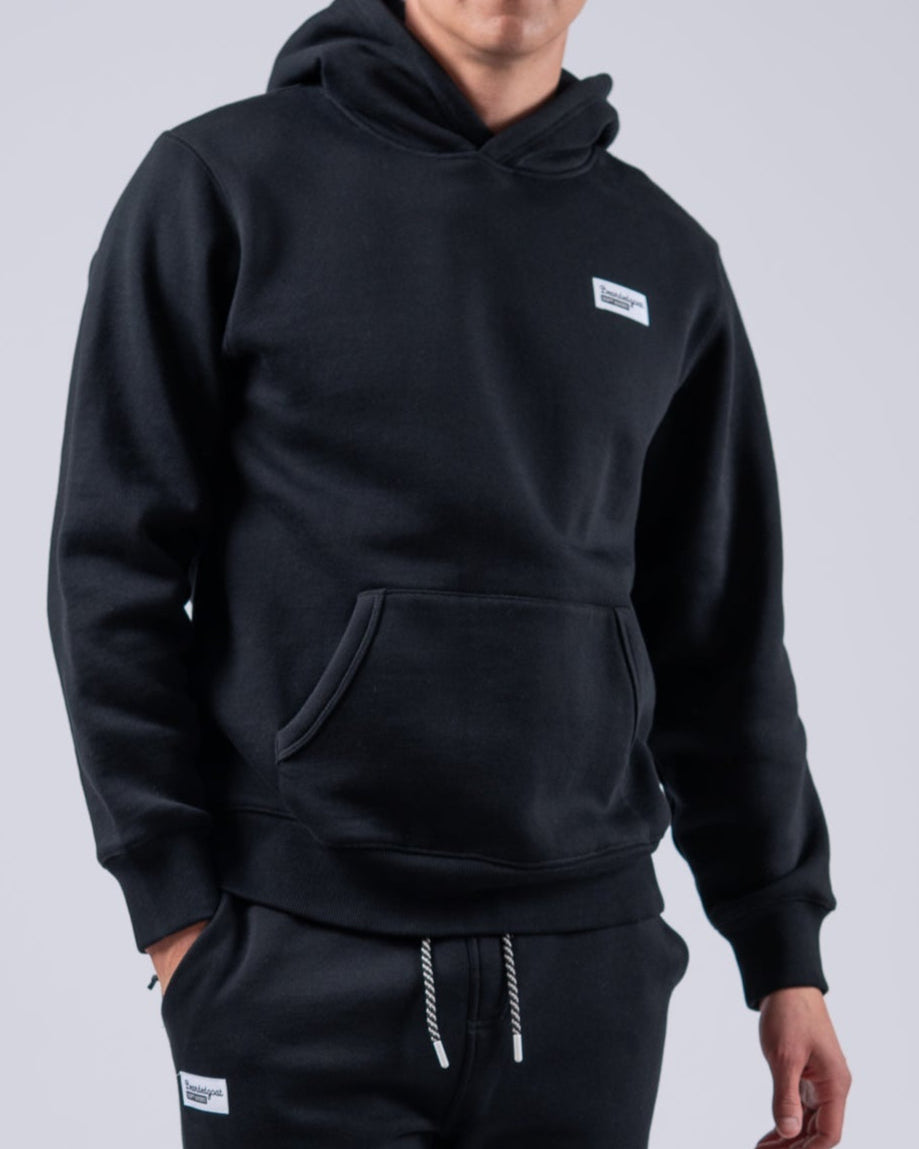 A person stands facing forward in a beardedgoat Mens Void Hoodie and matching black sweatpants with white sneakers, ideal for post-ride recovery, set against a plain light gray background.