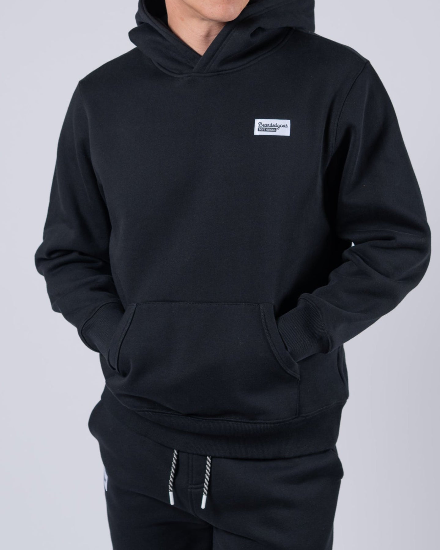 A young man with light brown hair wears the beardedgoat Mens Void Hoodie, made from heavyweight cotton fleece, paired with matching pants. He stands against a plain light gray background, hands in his hoodie pocket and looking slightly to the side.