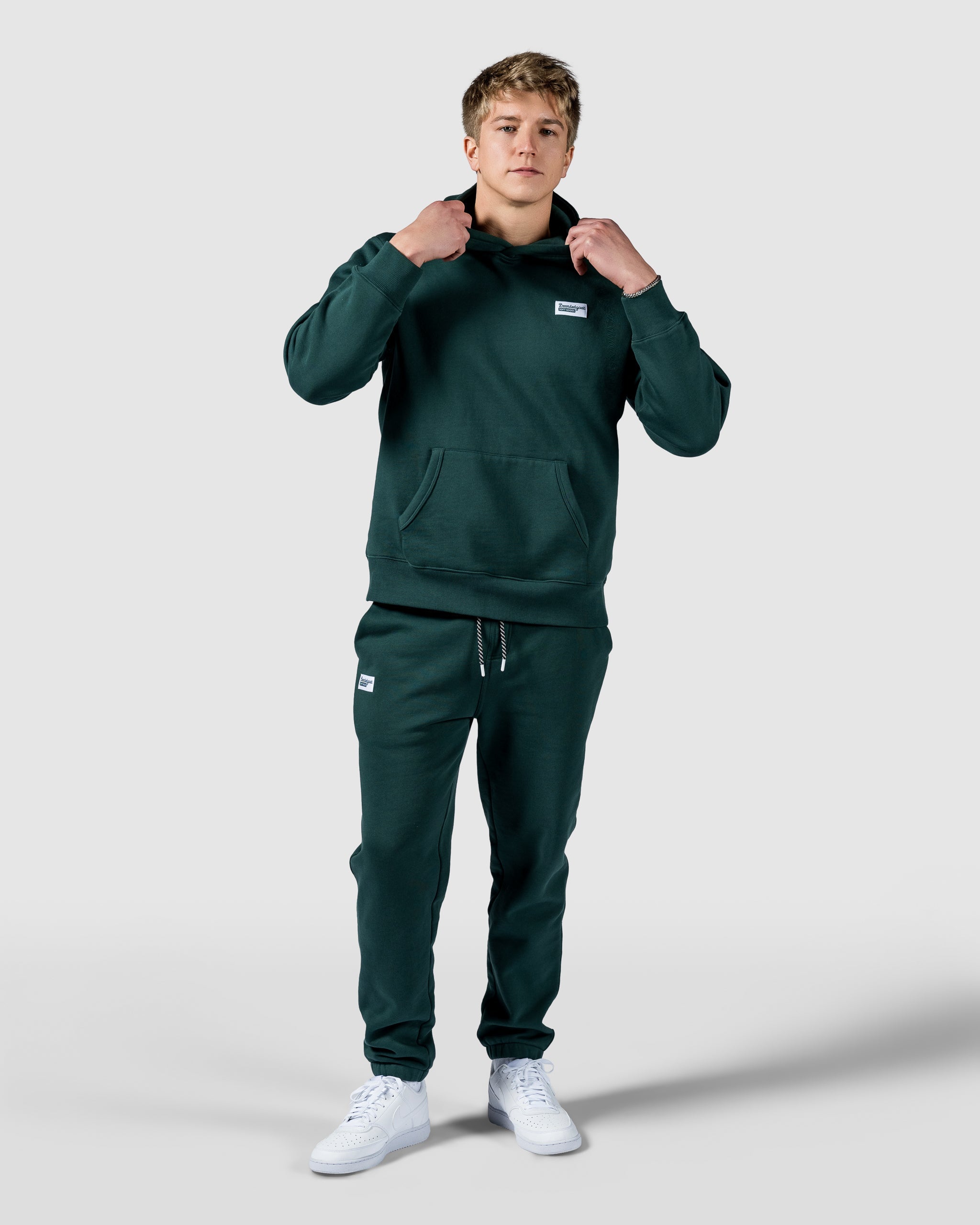A person stands against a plain background, adjusting the hood of a beardedgoat Mens Void Hoodie in dark green heavyweight cotton fleece with matching joggers and white sneakers, looking forward and ready for post-ride recovery.