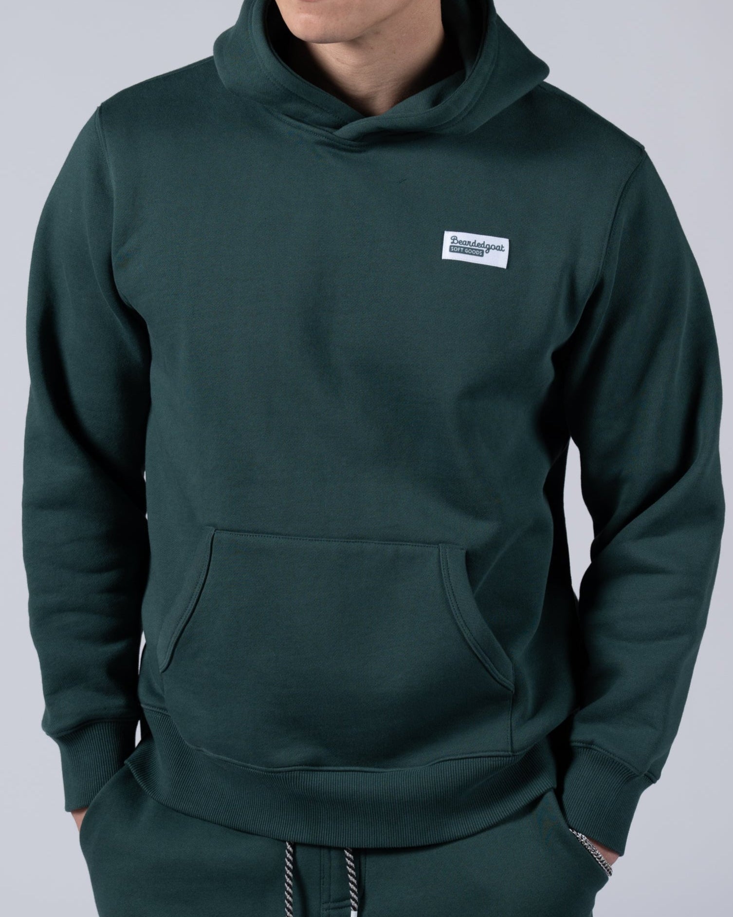 A young man wearing the beardedgoat Mens Void Hoodie in dark green, made from heavyweight cotton fleece, stands against a light gray background with hands in the hoodie pocket—ideal for post-ride recovery.