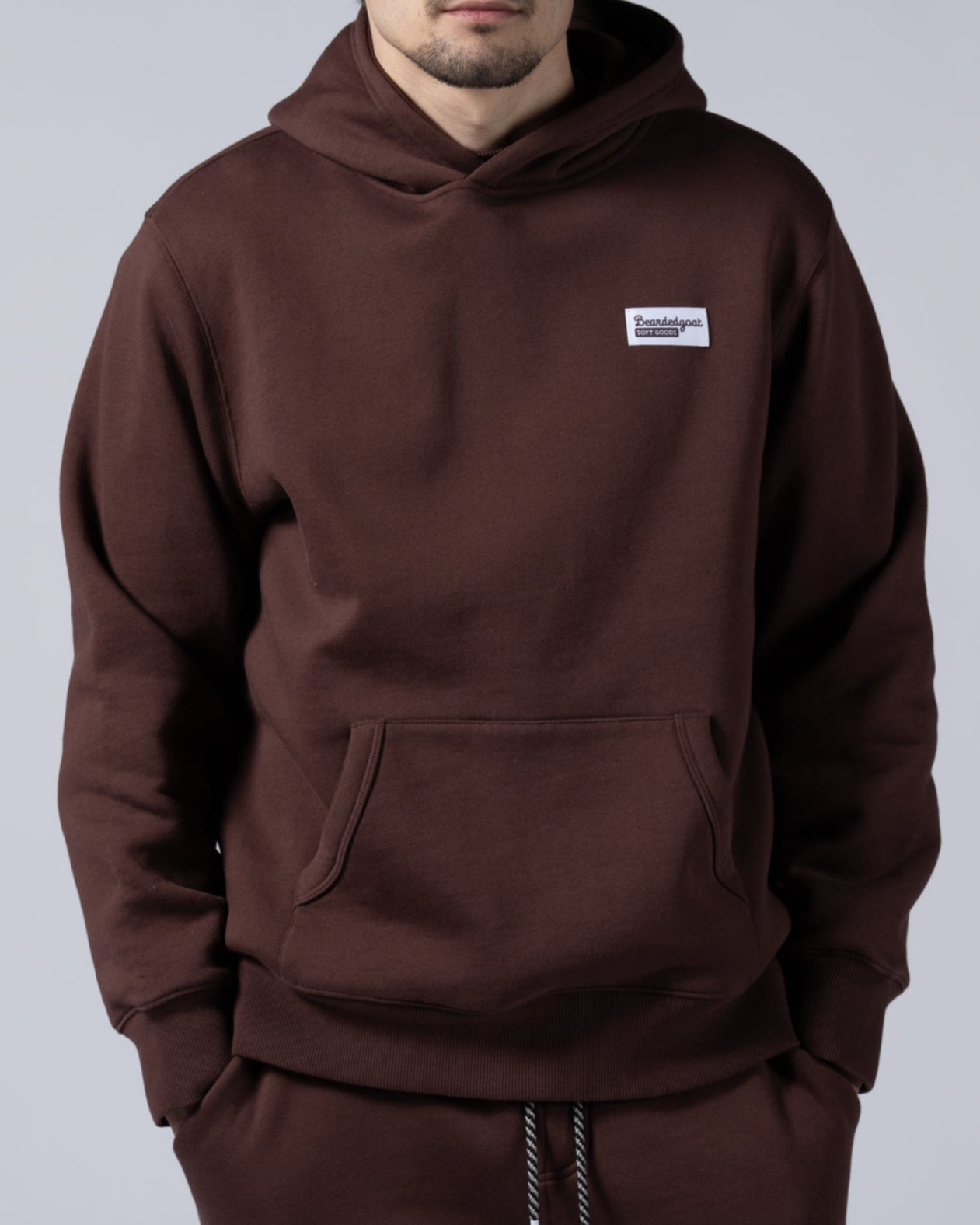 A man with short dark hair wears the beardedgoat Mens Void Hoodie in heavyweight cotton fleece, standing against a plain light gray background with his hands in his pockets—the ideal post-ride recovery look.