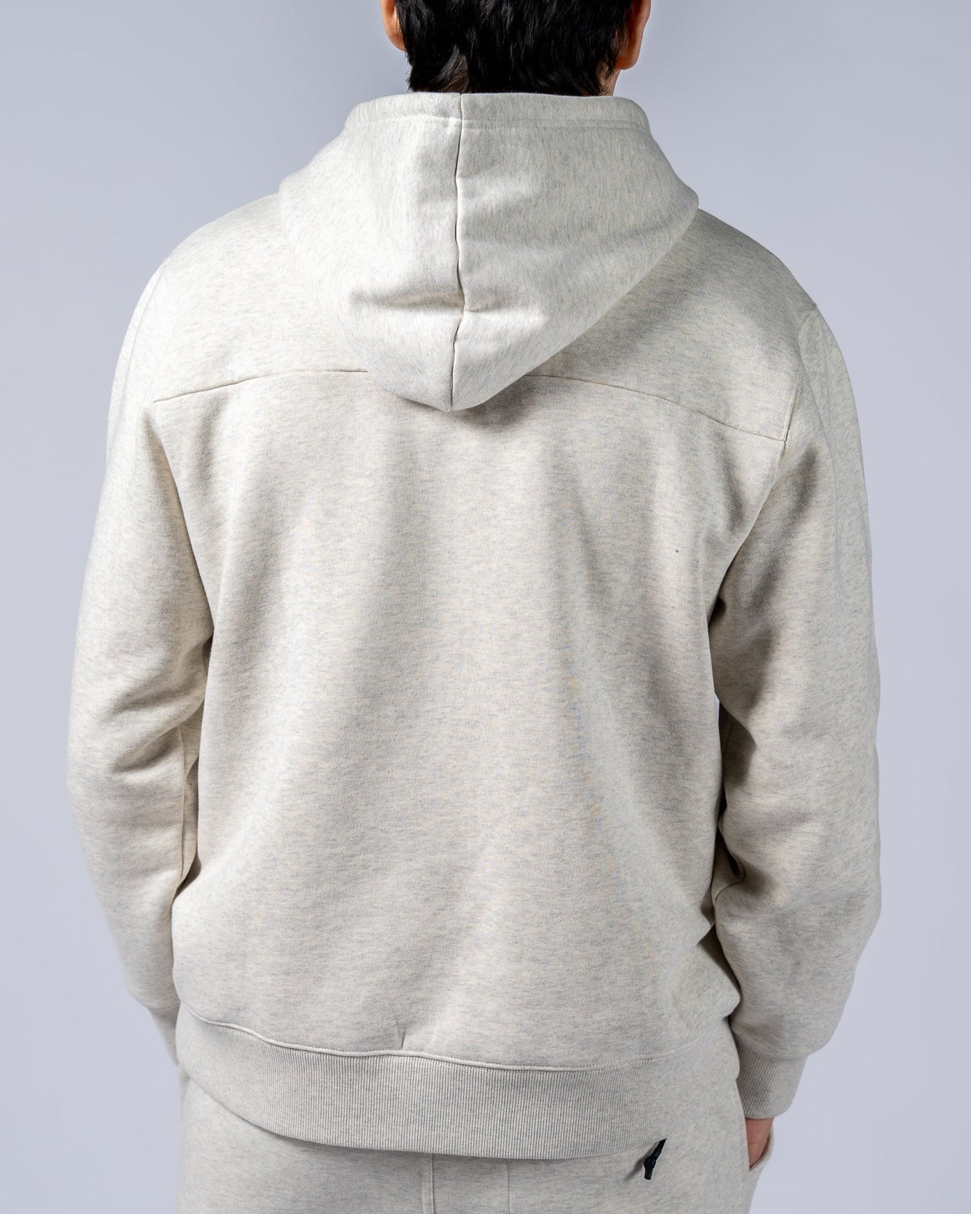 A man with dark hair stands with his back to the camera in a beardedgoat Mens Void Hoodie and matching sweatpants, made from heavyweight cotton fleece—ideal for post-ride recovery—against a plain light gray background.