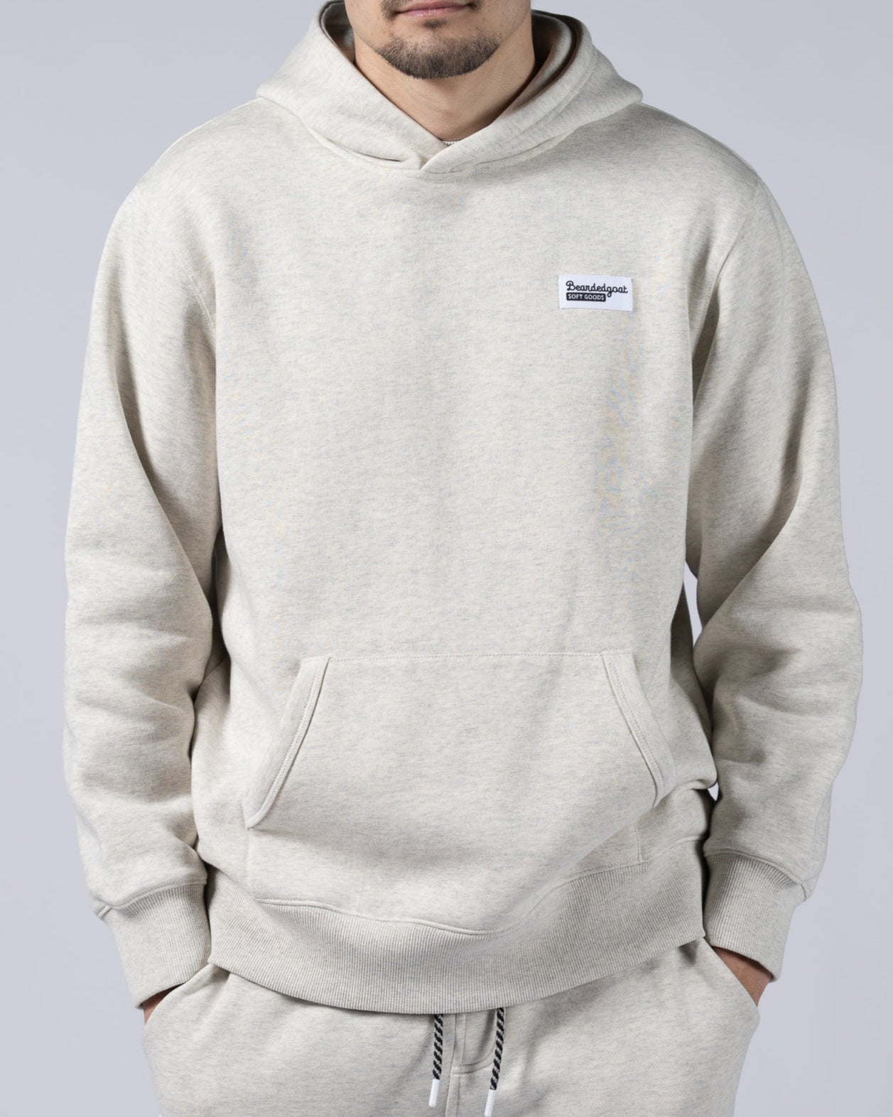 A man with short dark hair wears the beardedgoat Mens Void Hoodie in light beige, made from heavyweight cotton fleece, standing against a plain light gray background—ideal for post-ride recovery.