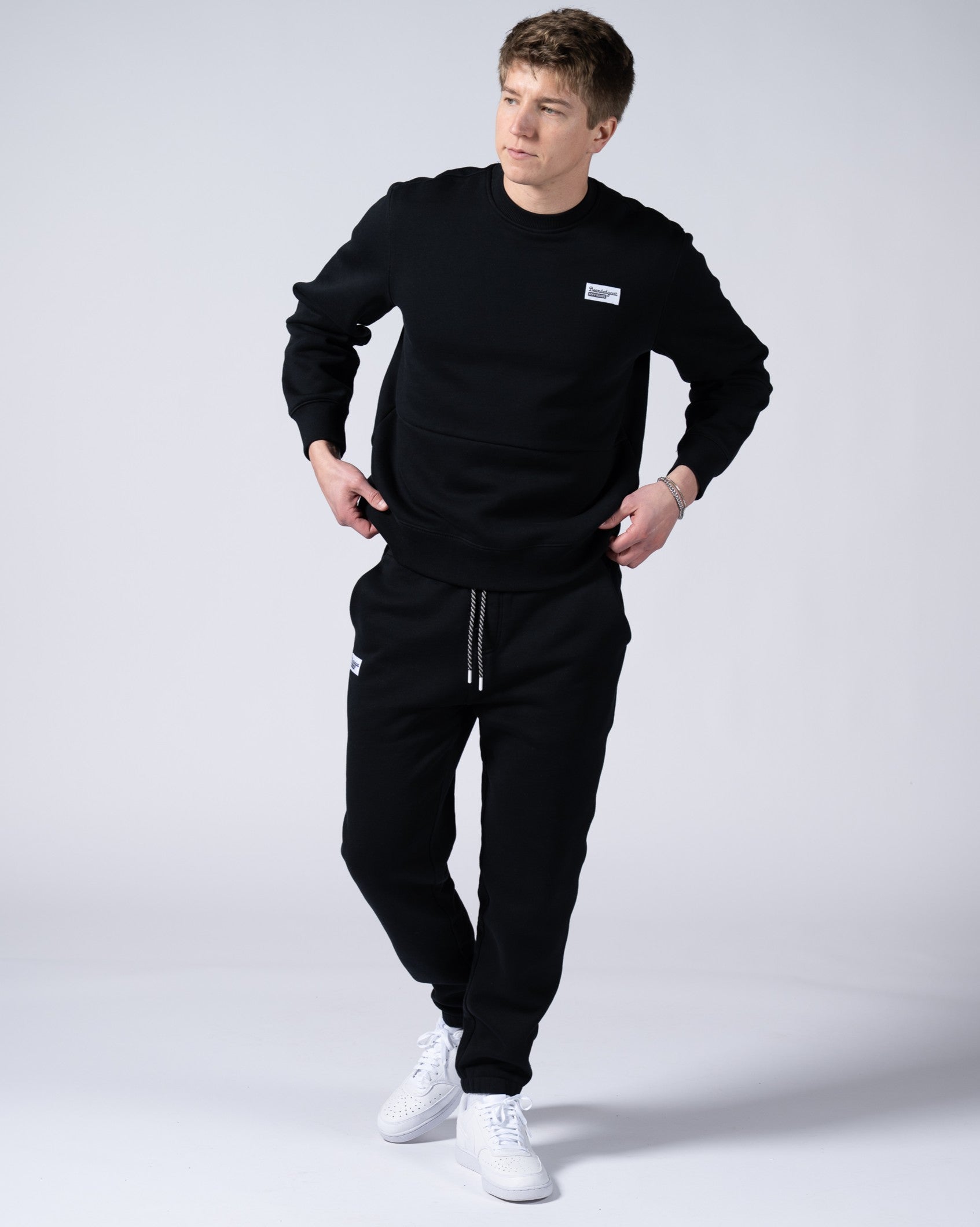 A man stands against a plain light background wearing beardedgoat Mens Void Sweatpants, paired with a black sweatshirt and white sneakers. He looks slightly to the side with one hand near his pocket.
