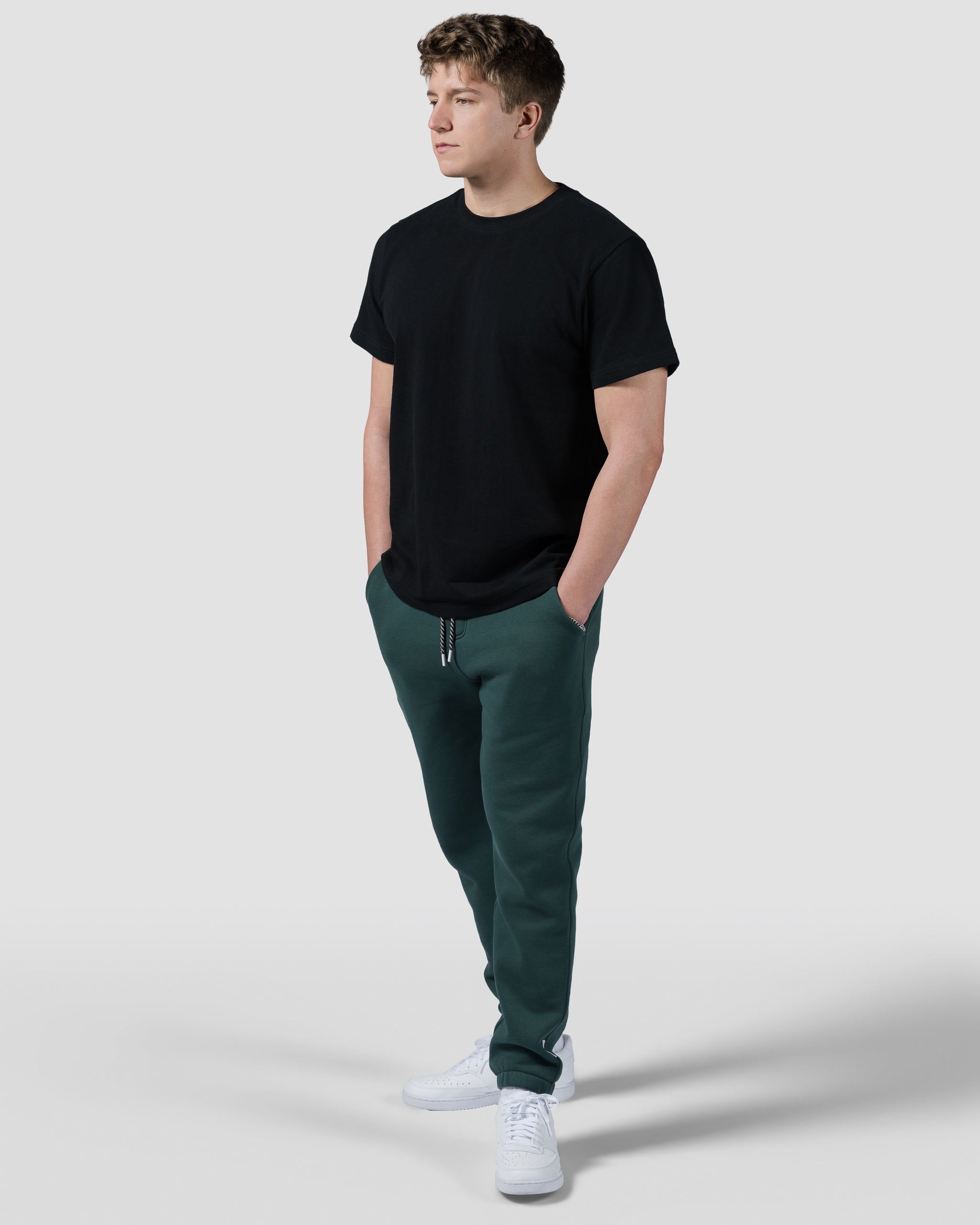 A young man stands with hands in pockets, wearing a black t-shirt, beardedgoat Mens Void Sweatpants in green, and white sneakers, set against a plain light gray background.