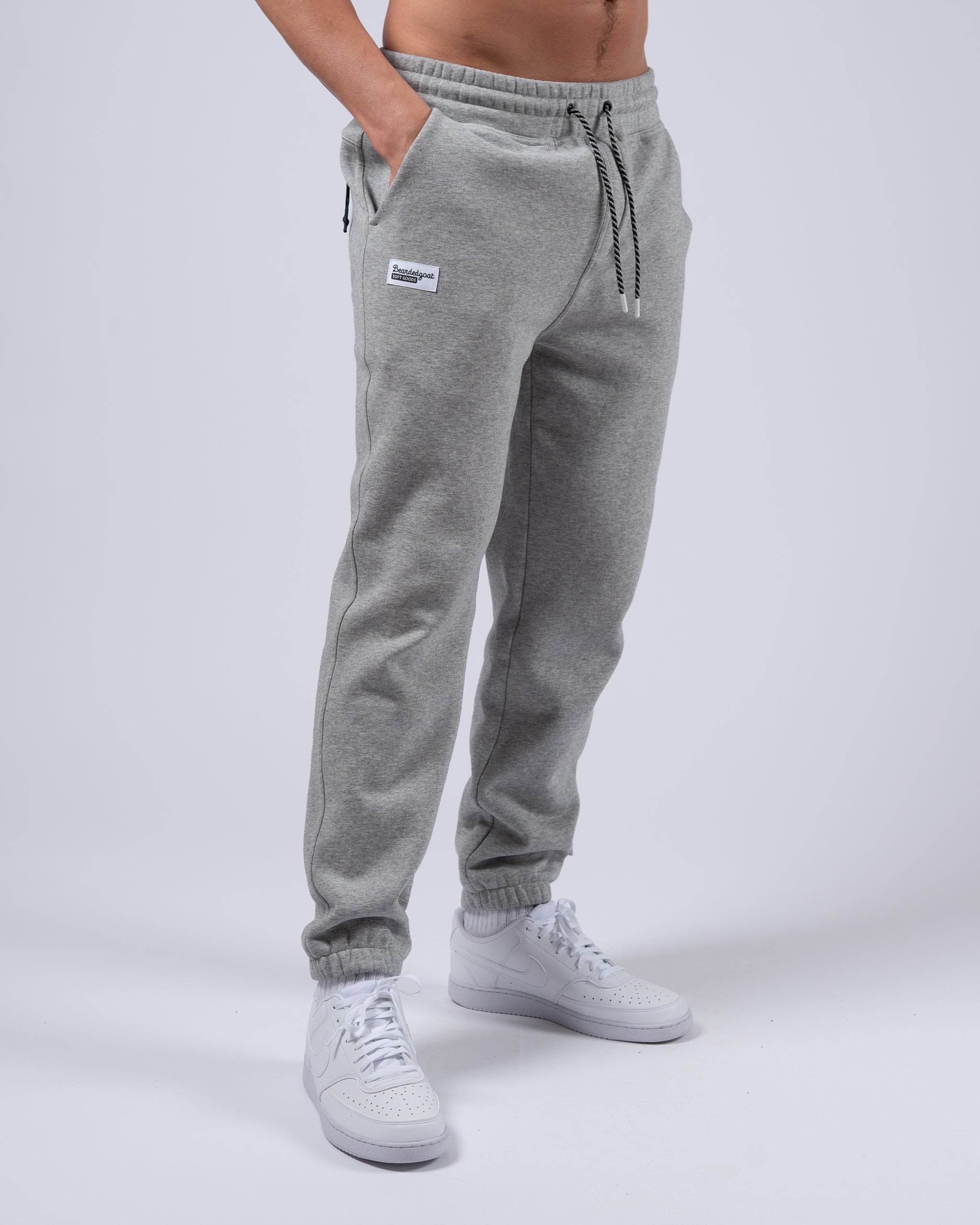 A person stands against a plain white background wearing beardedgoat Men's Void Sweatpants in light grey, featuring a drawstring waistband and zipper back pocket, paired with white athletic sneakers and hands in pockets.