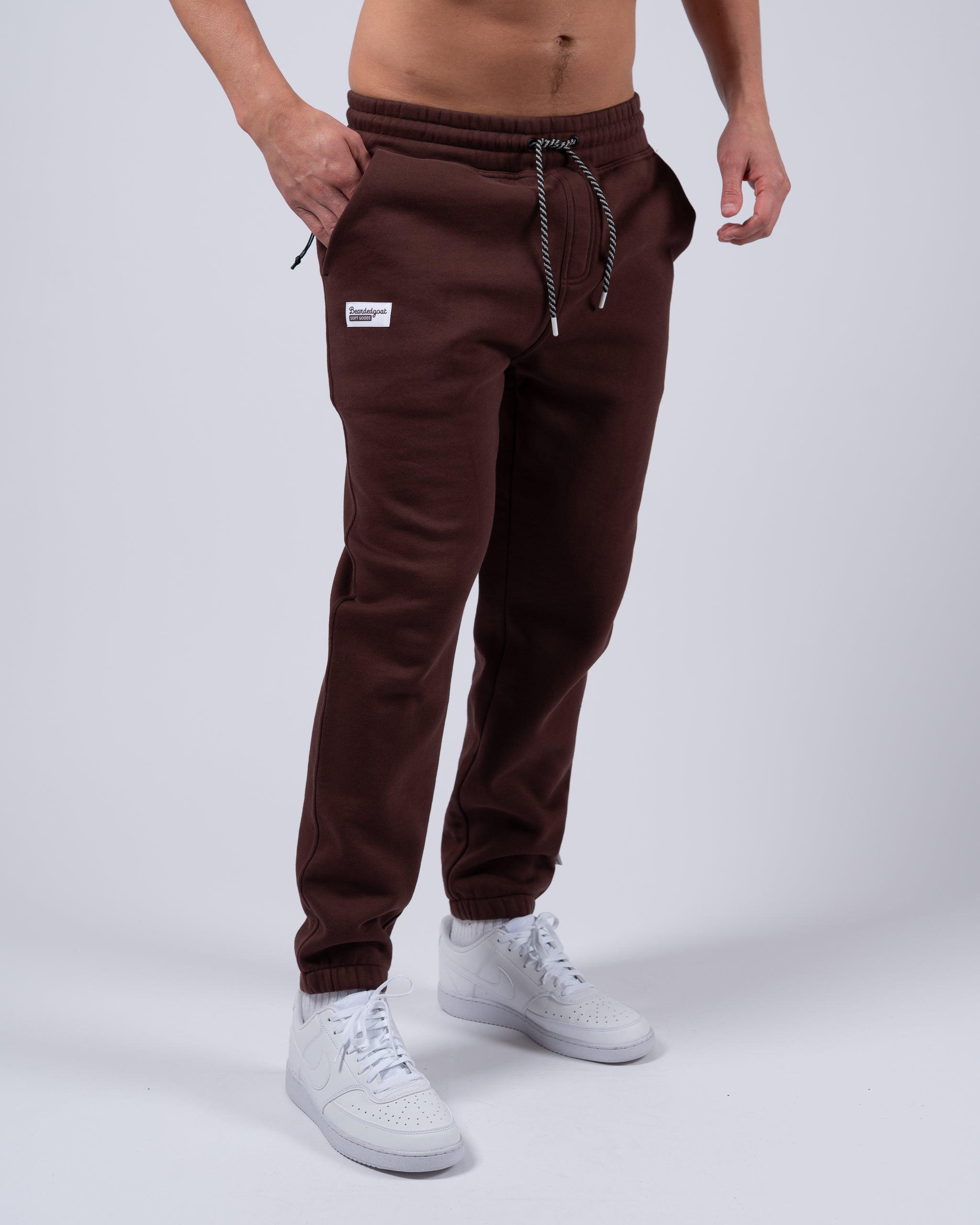 A person wears beardedgoat Men's Void Sweatpants in brown, featuring a drawstring waist, white patch, and zippered back pocket, paired with white sneakers, against a plain light gray background. Only the lower body is shown.