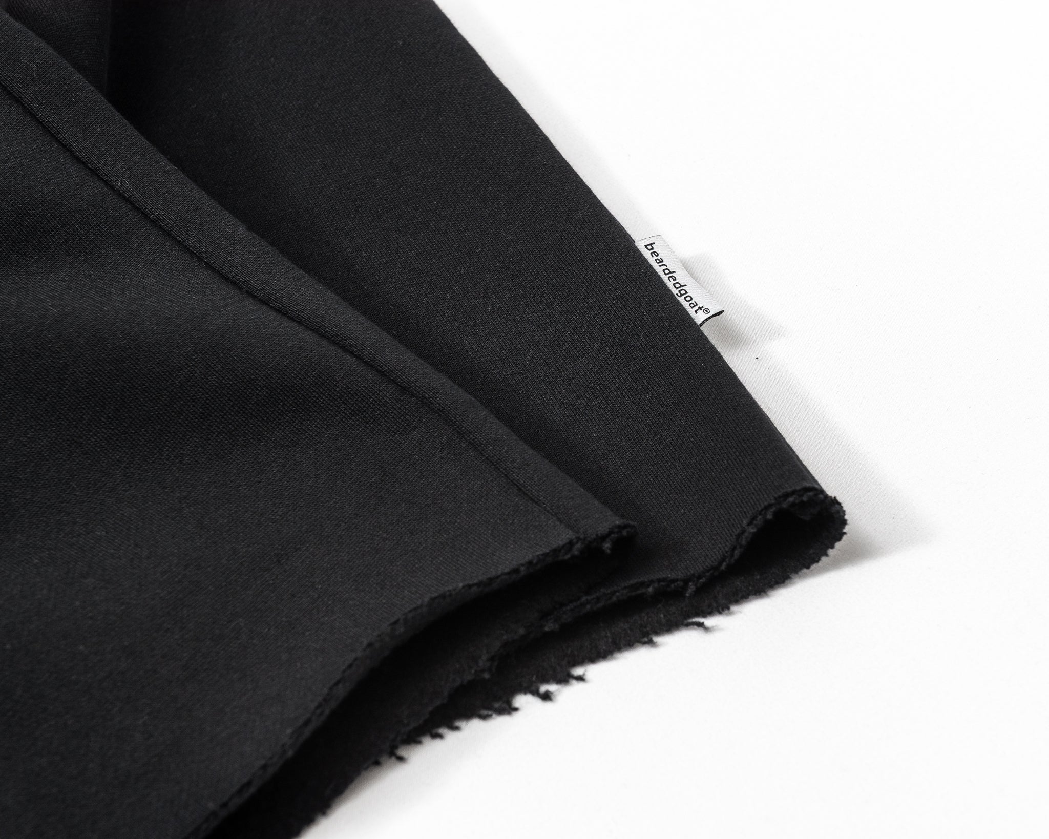 A close-up of Womens Overnight Sweatpants by beardedgoat shows black fabric with a raw, unfinished edge like raw-hem sweats and a small white bewusstkraft tag, displayed on a white surface.