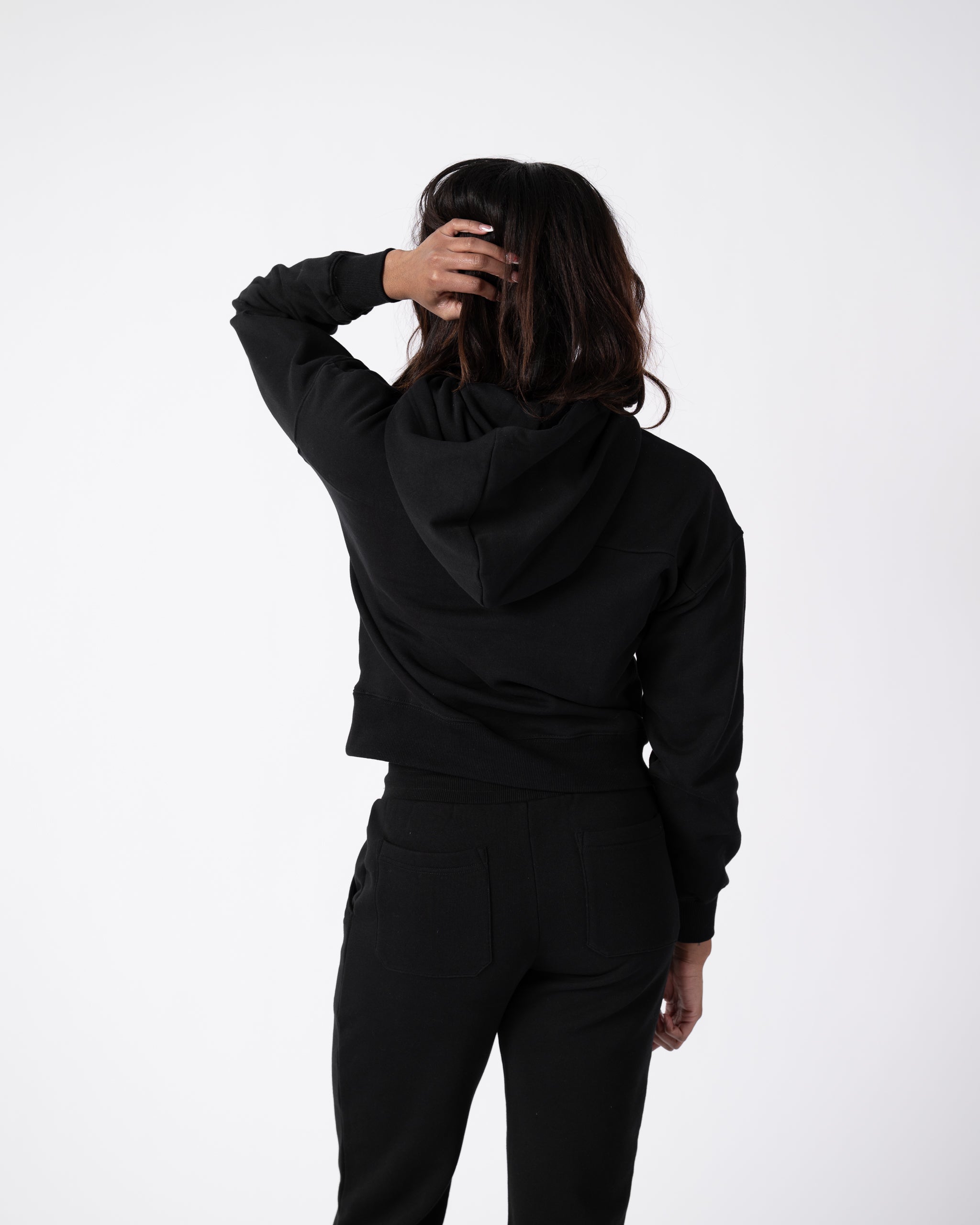 A person faces away against a white background, wearing beardedgoat Womens Overnight Sweatpants and a black hoodie with beardedgoat script embroidery, one hand lifted to the back of their head.