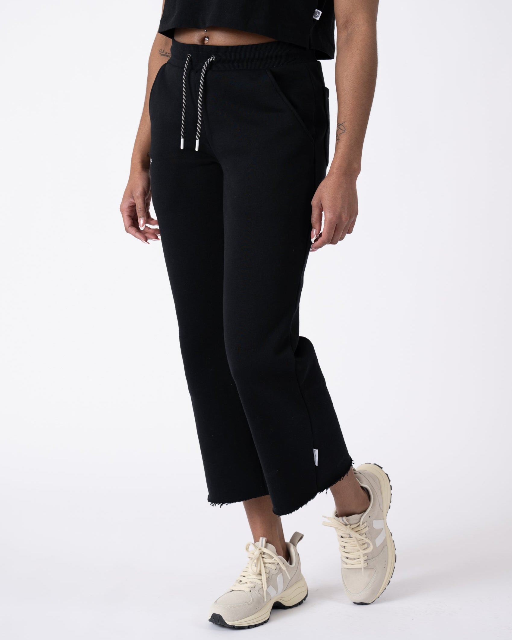 A model wears the beardedgoat Women’s Overnight Sweatpant in heavyweight black cotton fleece with a drawstring waist and raw hems, styled with a black top and beige sneakers, set against a plain white background.