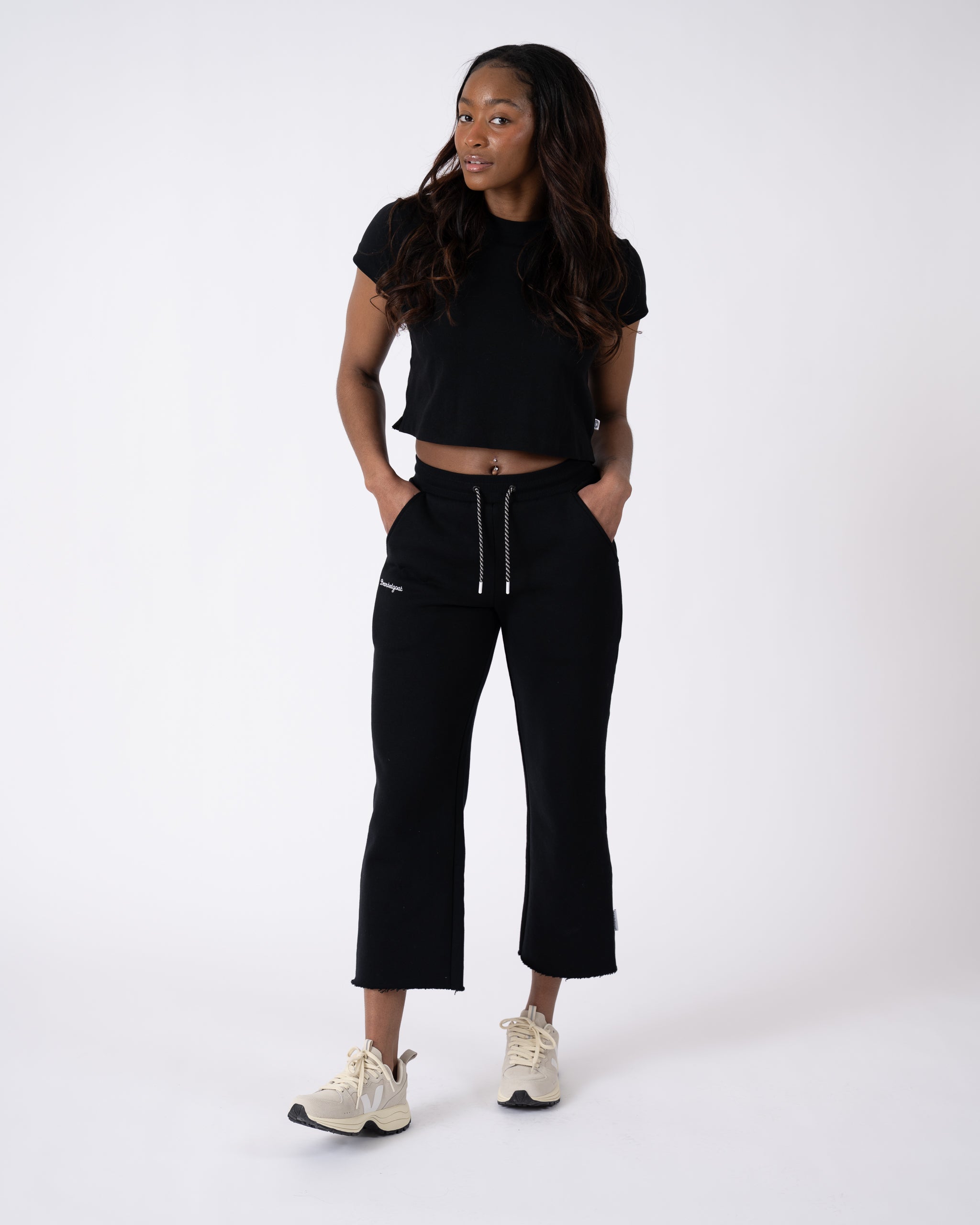 A woman stands against a white background in a black cropped tee, beardedgoat Womens Overnight Sweatpants with script embroidery, and white sneakers, hands in her pockets as she gazes to the side.