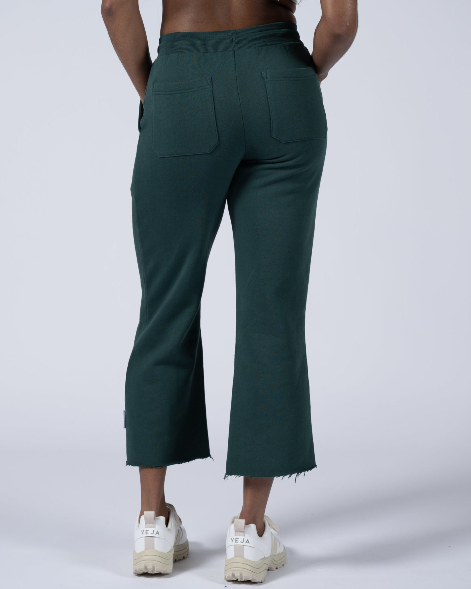 A person is seen from behind wearing beardedgoat Women’s Overnight Sweatpants in dark green with frayed hems and white Veja sneakers against a plain, light background.