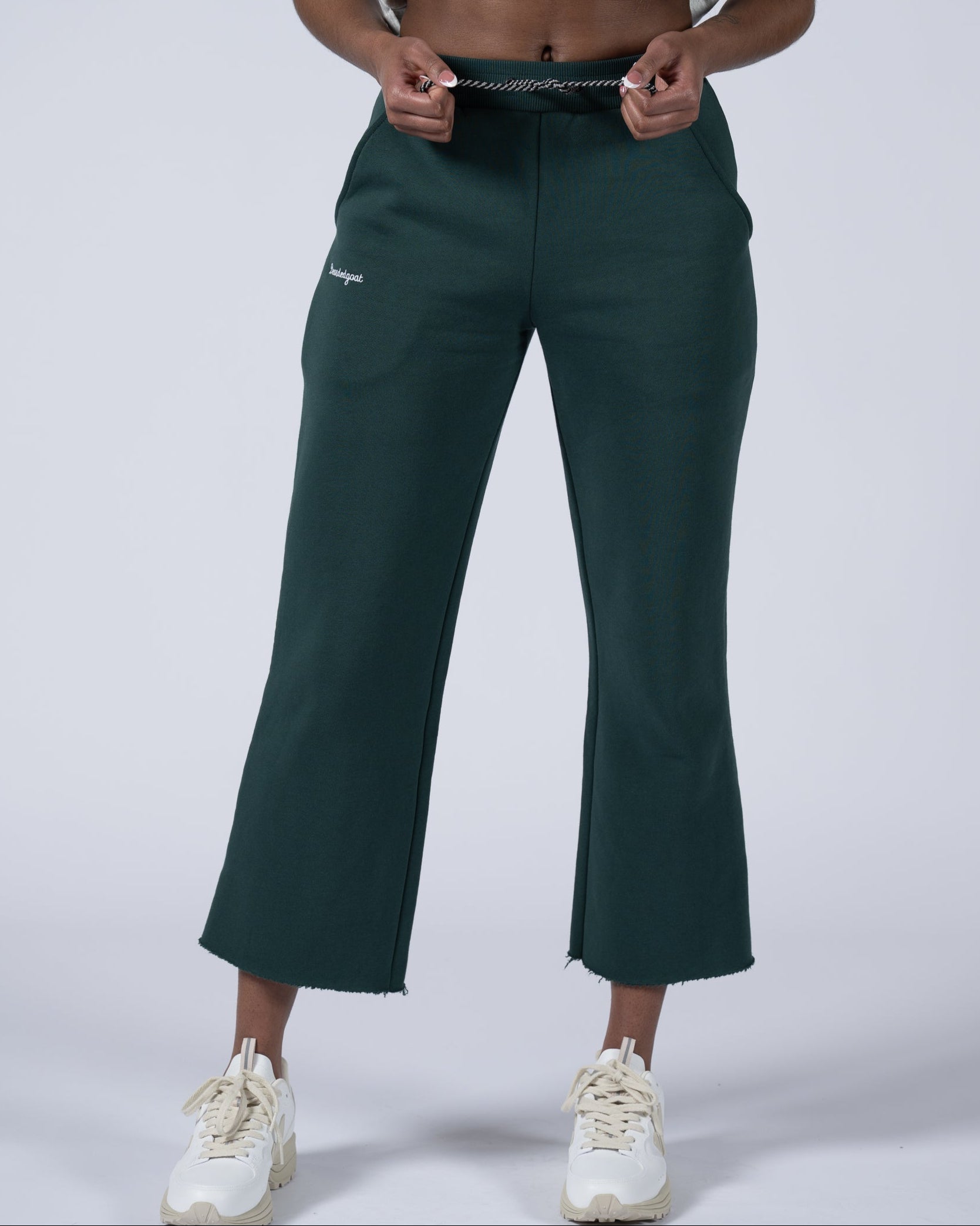 A person wearing beardedgoat’s Women’s Overnight Sweatpant in dark green—a wide-leg, heavyweight cotton fleece style with a drawstring waist and frayed hems—paired with white sneakers, stands against a plain light backdrop.