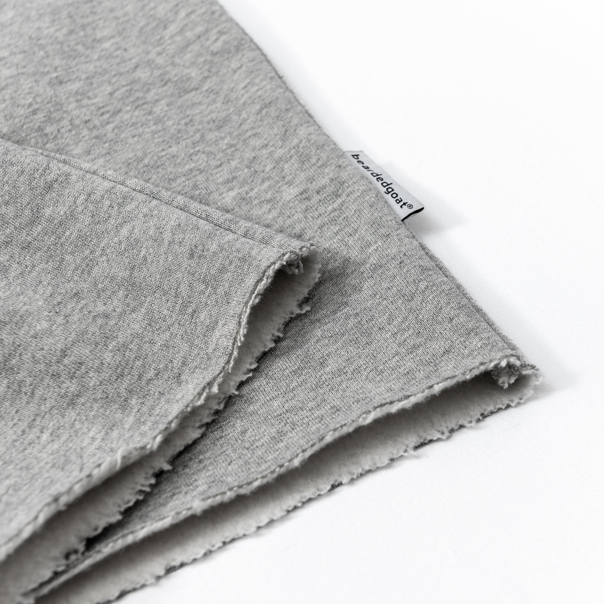 A close-up of light gray Womens Overnight Sweatpants by beardedgoat with raw hems and a small white tag by the seam. The pants lie flat on a white background, revealing subtle beardedgoat script embroidery.