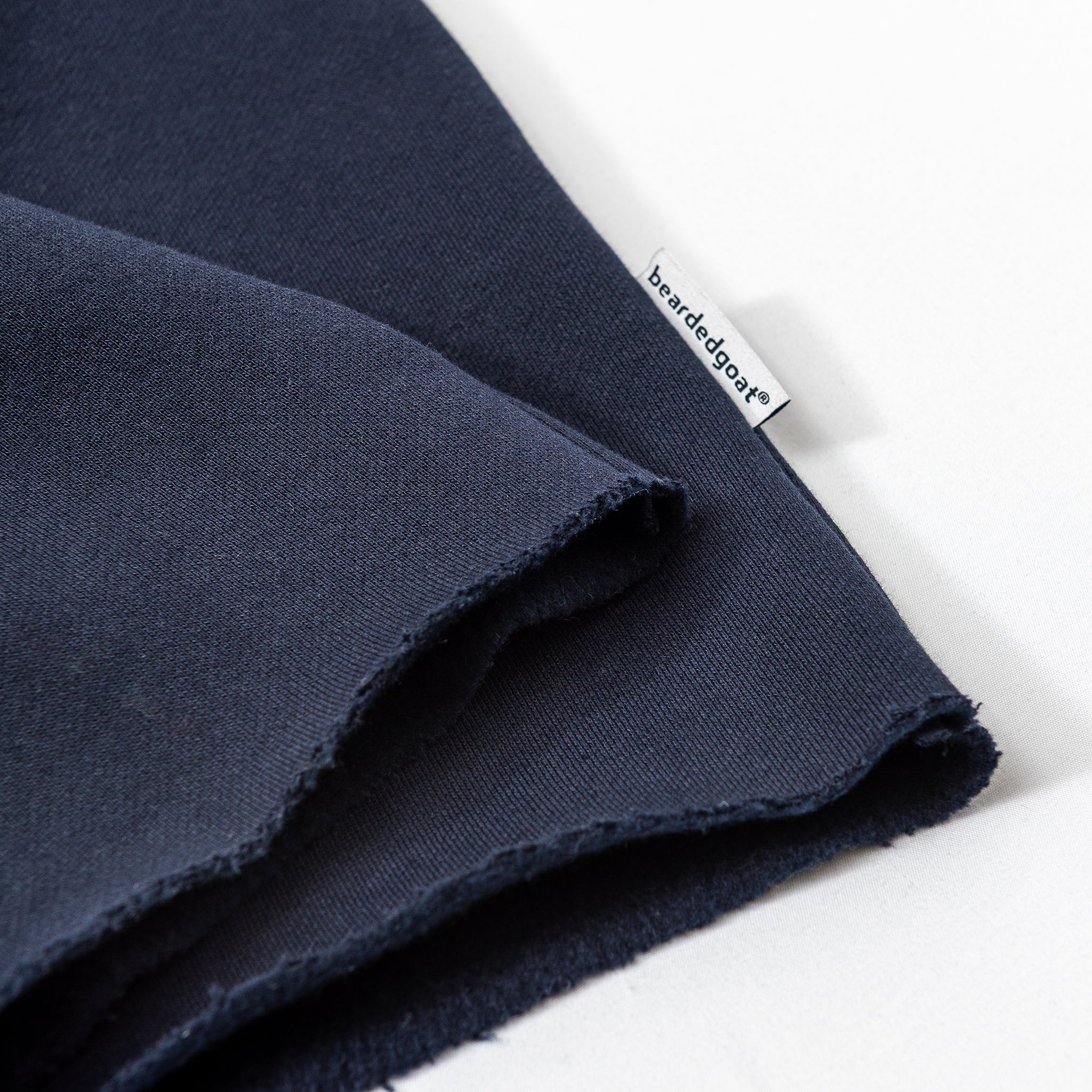 Close-up of two overlapping pieces of dark navy blue Womens Overnight Sweatpants by beardedgoat, featuring a small white tag with signature beardedgoat script embroidery, placed on a white surface.