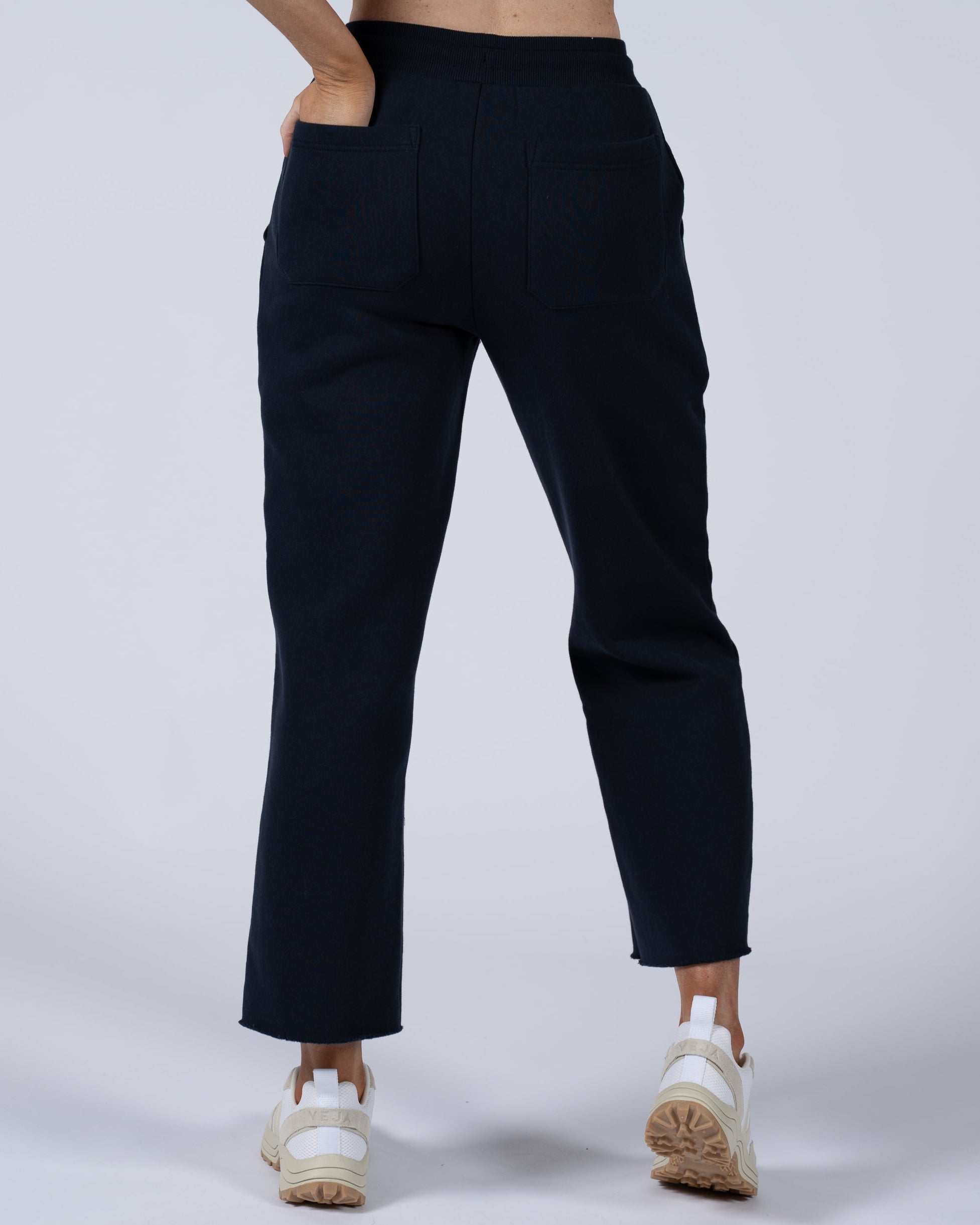 A person is shown from the waist down wearing beardedgoat Women’s Overnight Sweatpants in black with back pockets and white sneakers, set against a plain light background that highlights this comfortable loungewear.