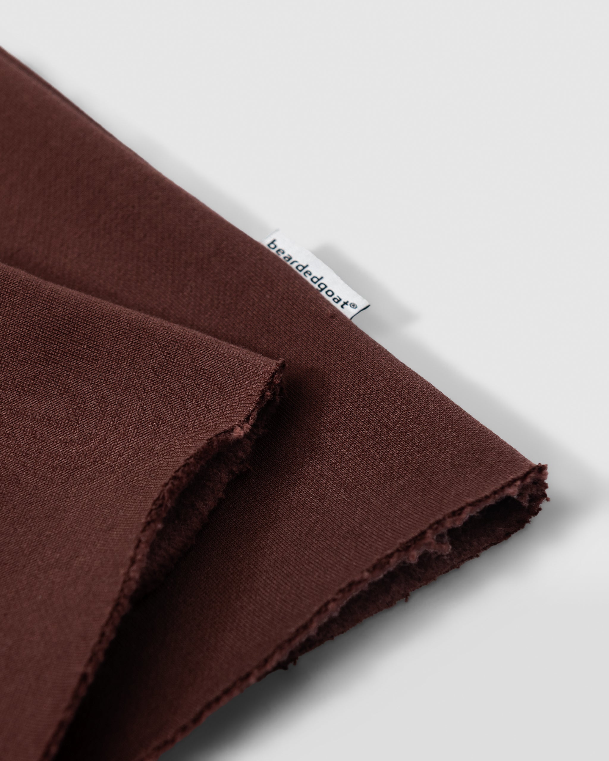 A close-up of two pieces of dark brown fabric—one folded over the other—shows a small white base de dose tag attached, highlighting the soft texture and unfinished edges of beardedgoat Womens Overnight Sweatpants.