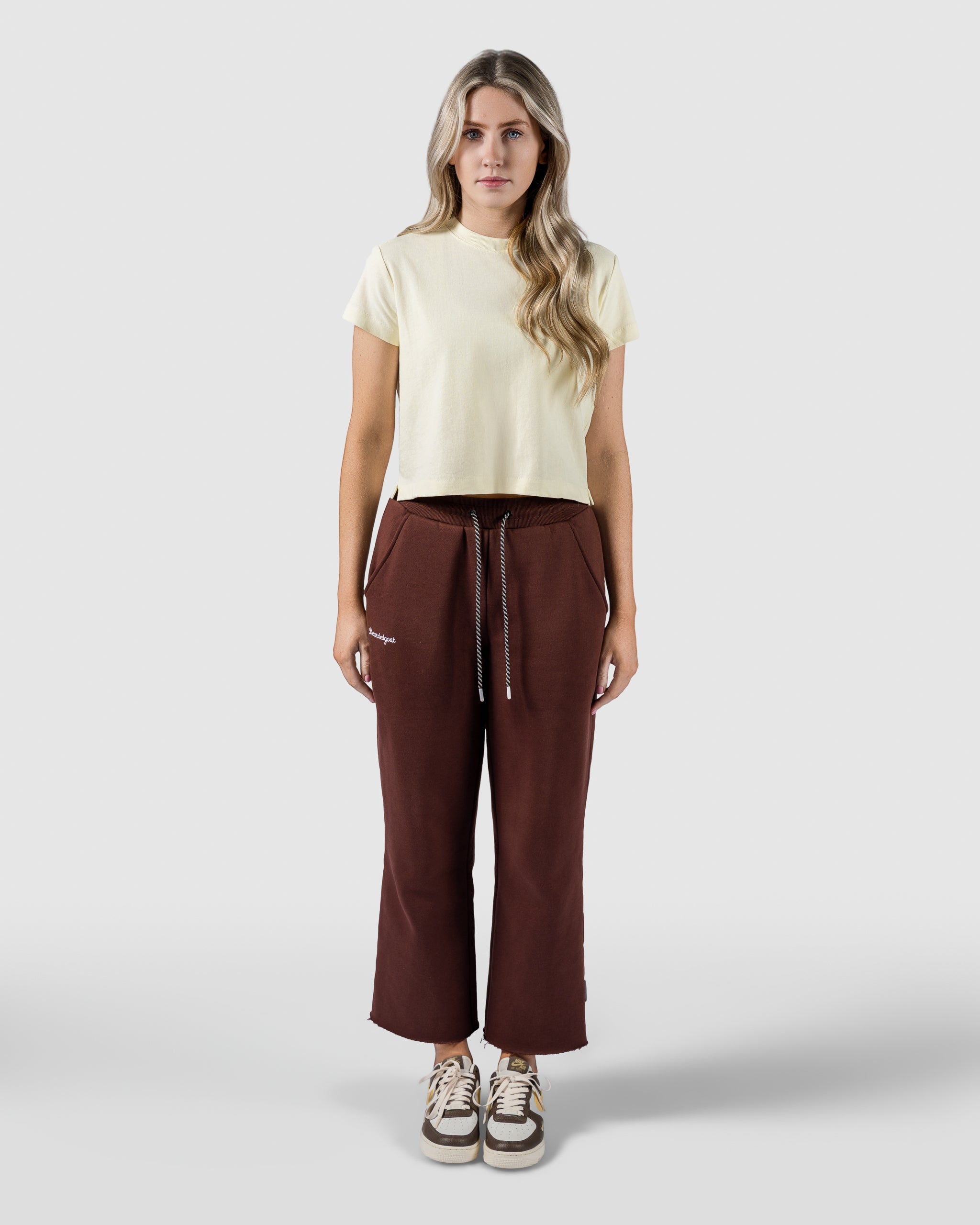 A woman with long, wavy blond hair faces forward wearing beardedgoat Womens Overnight Sweatpants, a light yellow t-shirt, and beige sneakers against a plain light gray background.