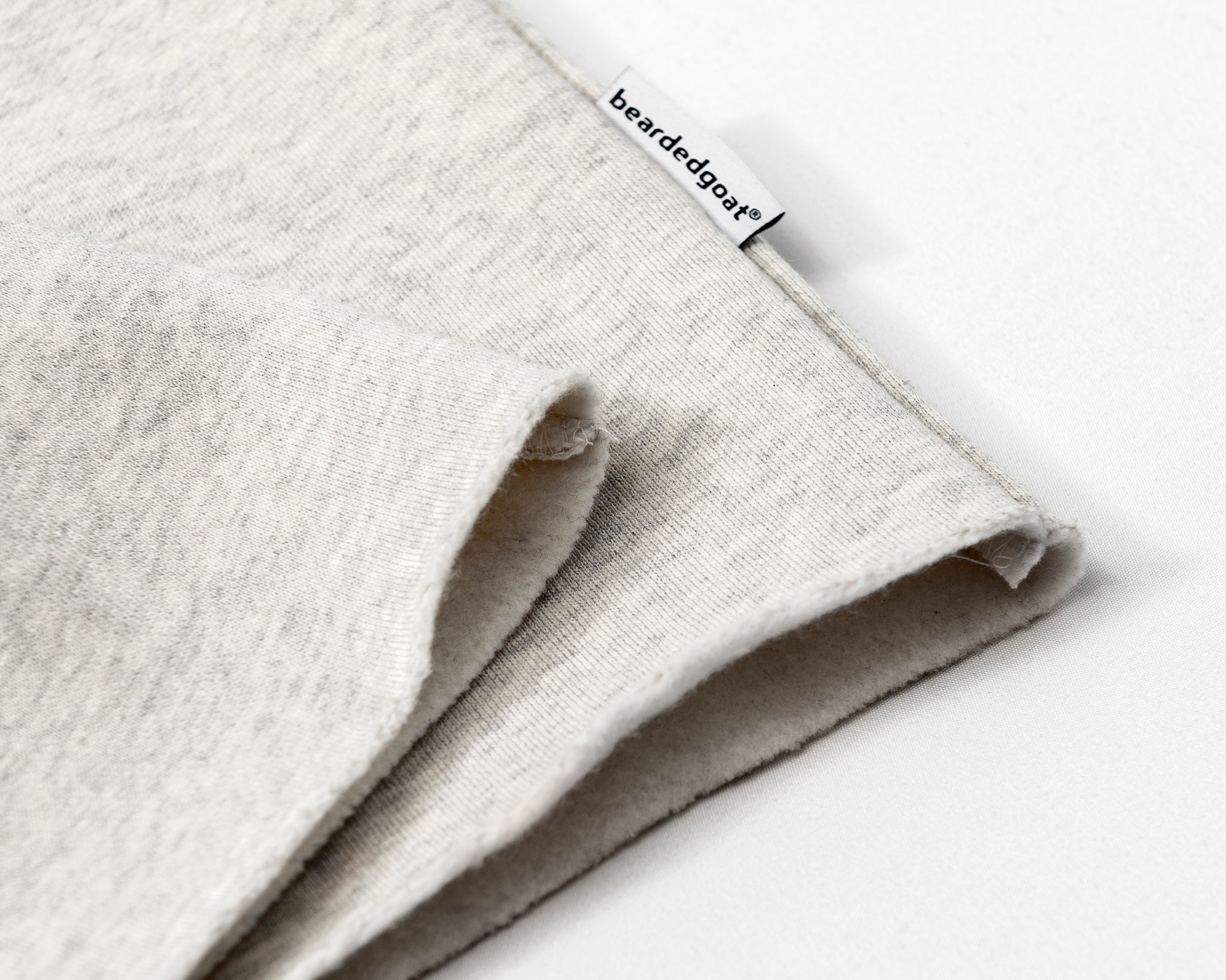 A close-up of light gray Womens Overnight Sweatpants by beardedgoat, featuring a raw hem and a small white beardedgoat tag on the edge, set against a plain white background.