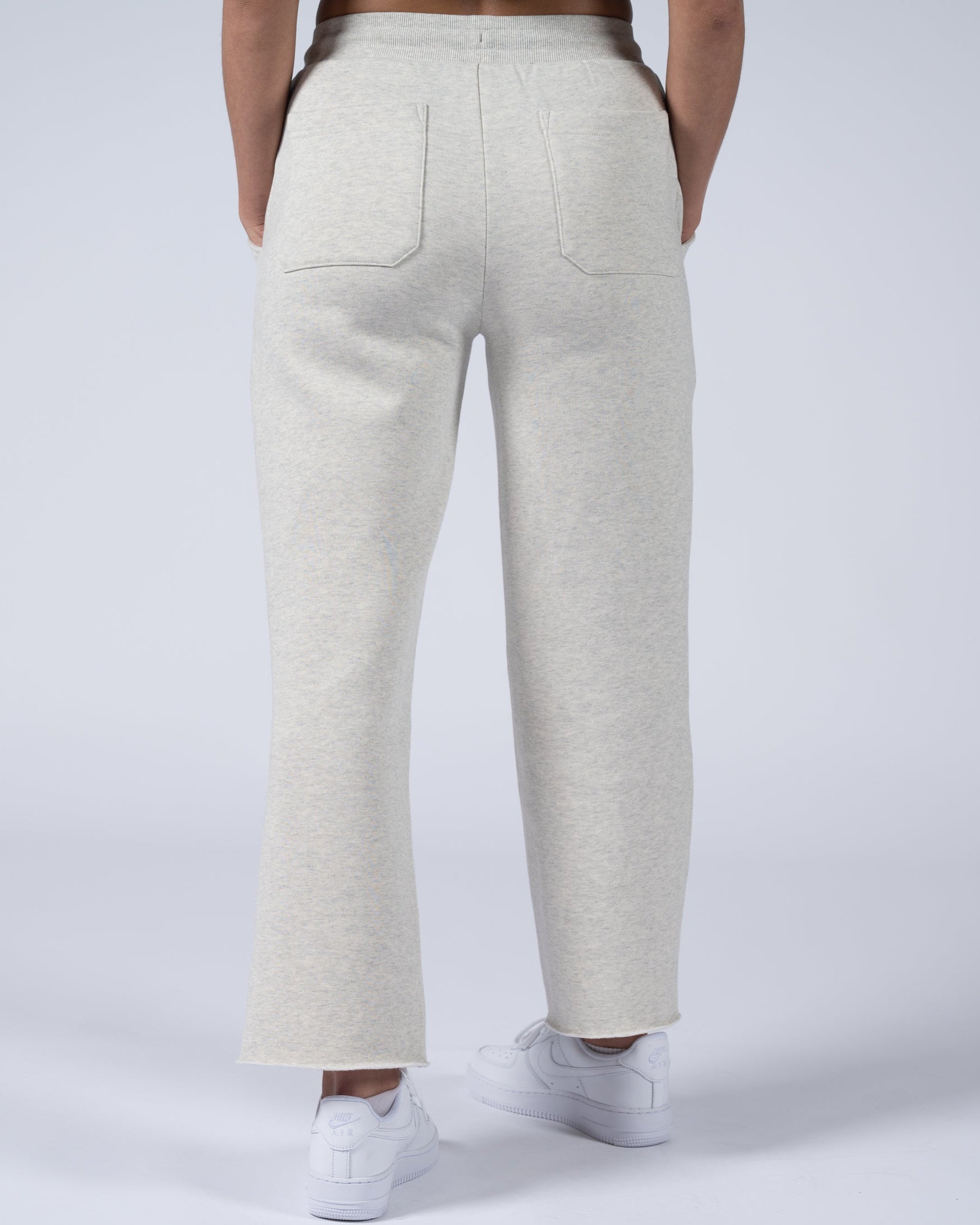 A person faces away from the camera, wearing beardedgoat’s Women’s Overnight Sweatpant in light gray heavyweight cotton fleece with two back pockets and a relaxed fit, paired with white sneakers. The background is plain and light.