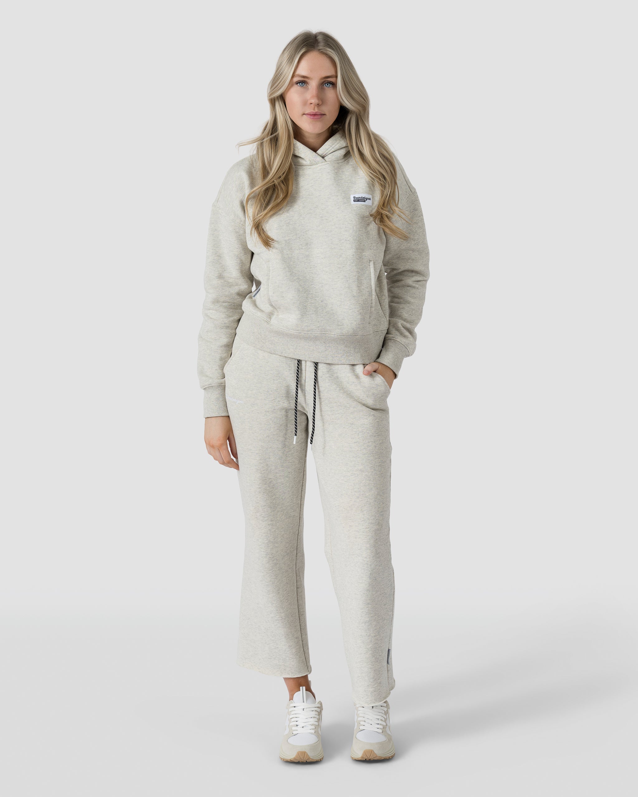 A woman with long, wavy blonde hair faces forward in beardedgoat Womens Overnight Sweatpants, light gray with script embroidery and raw hems. She has her left hand in her pocket, wears white sneakers, and stands against a plain white background.