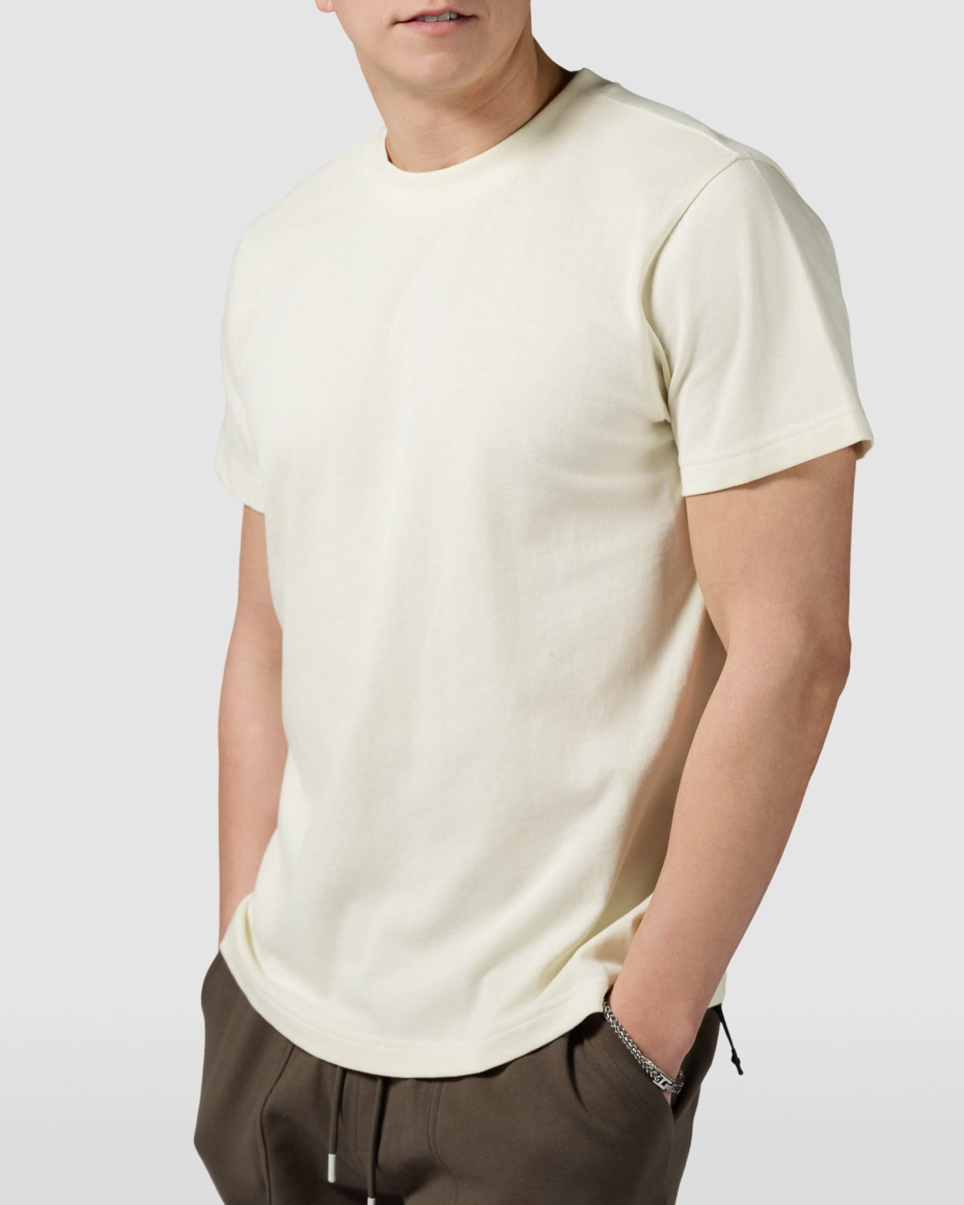 A young man with short light brown hair stands against a plain background, wearing the BEARDEDGOAT Mens Valley Tee in cream and dark brown shorts, with his hands in his shorts pockets.