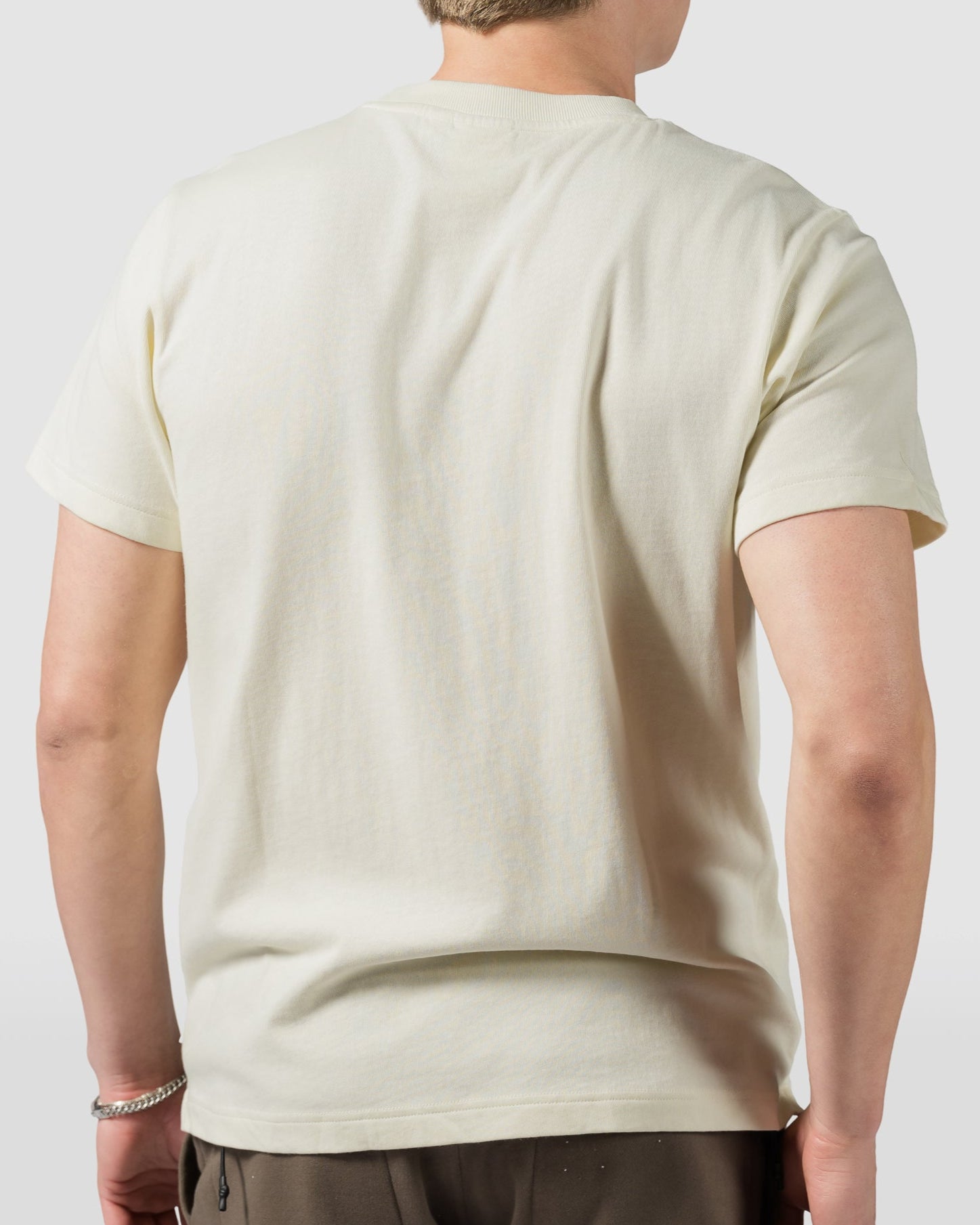 A person with short light brown hair, seen from behind, wears a light beige BEARDEDGOAT Mens Valley Tee and brown pants against a plain, light background.
