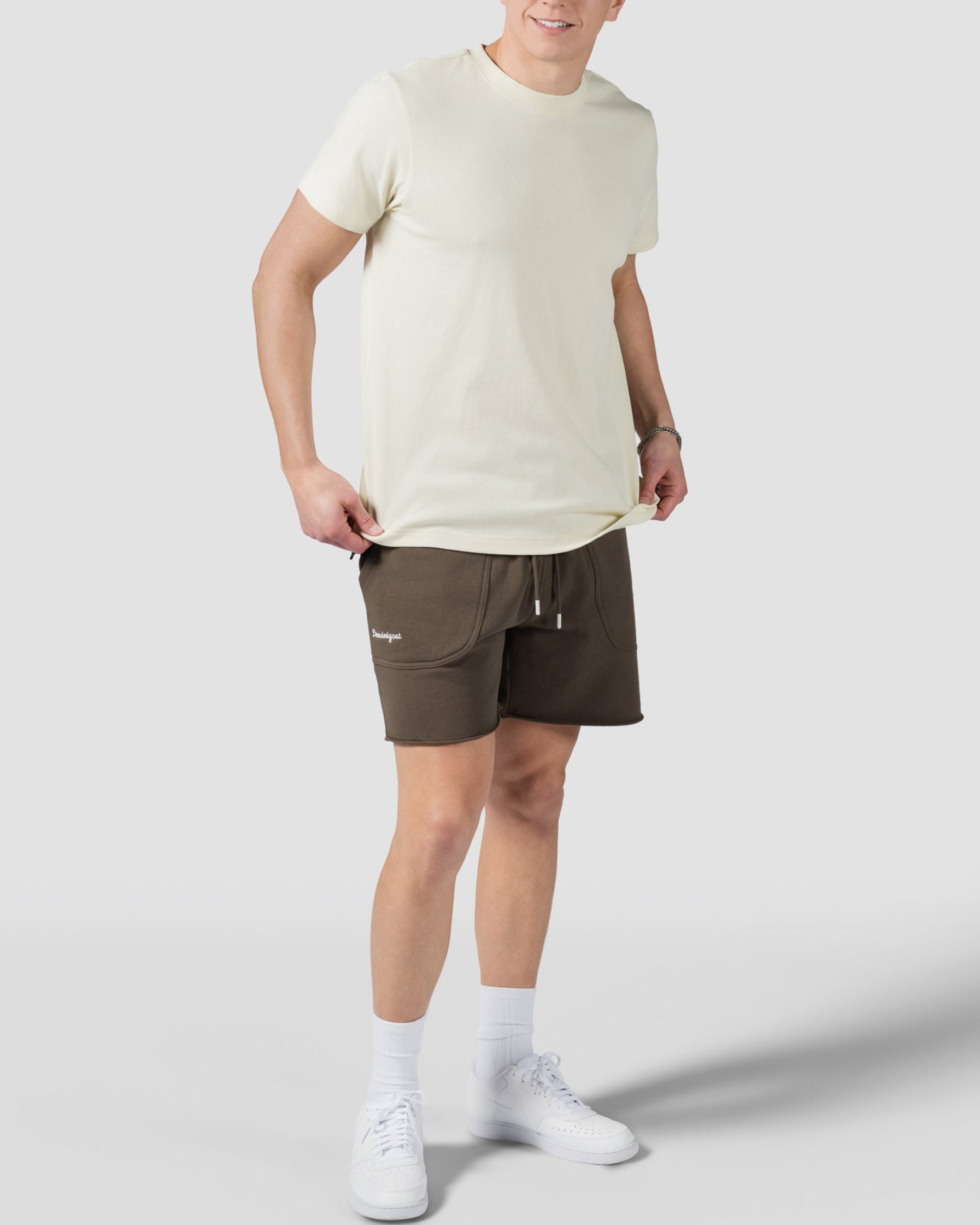 A young man smiles, wearing the BEARDEDGOAT Mens Valley Tee in cream, dark olive drawstring shorts, white crew socks, and white sneakers, standing against a plain light gray background.