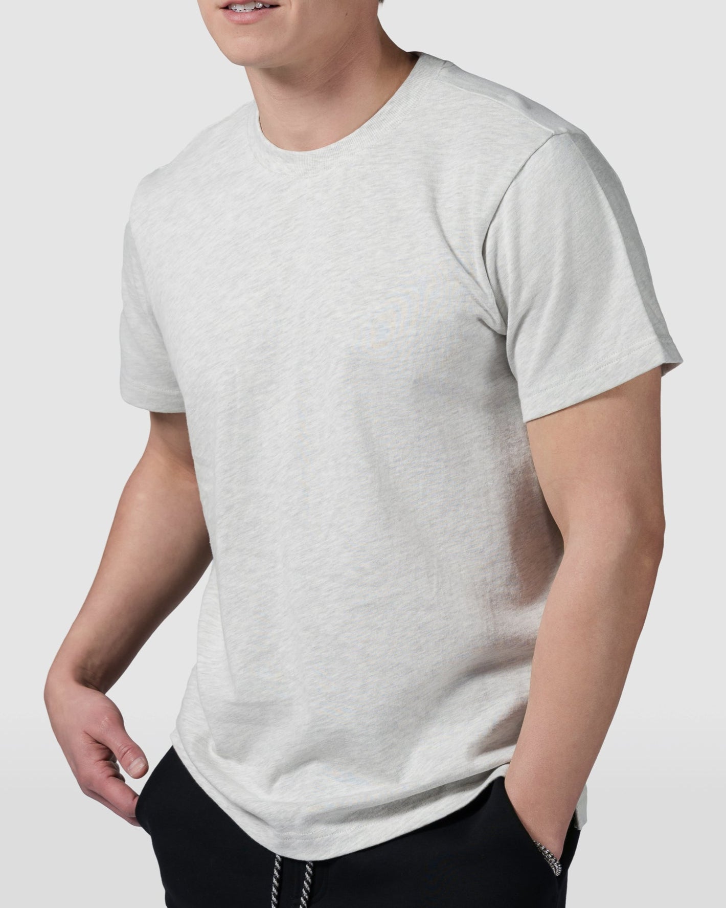 A young man wears a light gray BEARDEDGOAT Mens Valley Tee and black pants, standing with hands in pockets against a plain, light gray background.