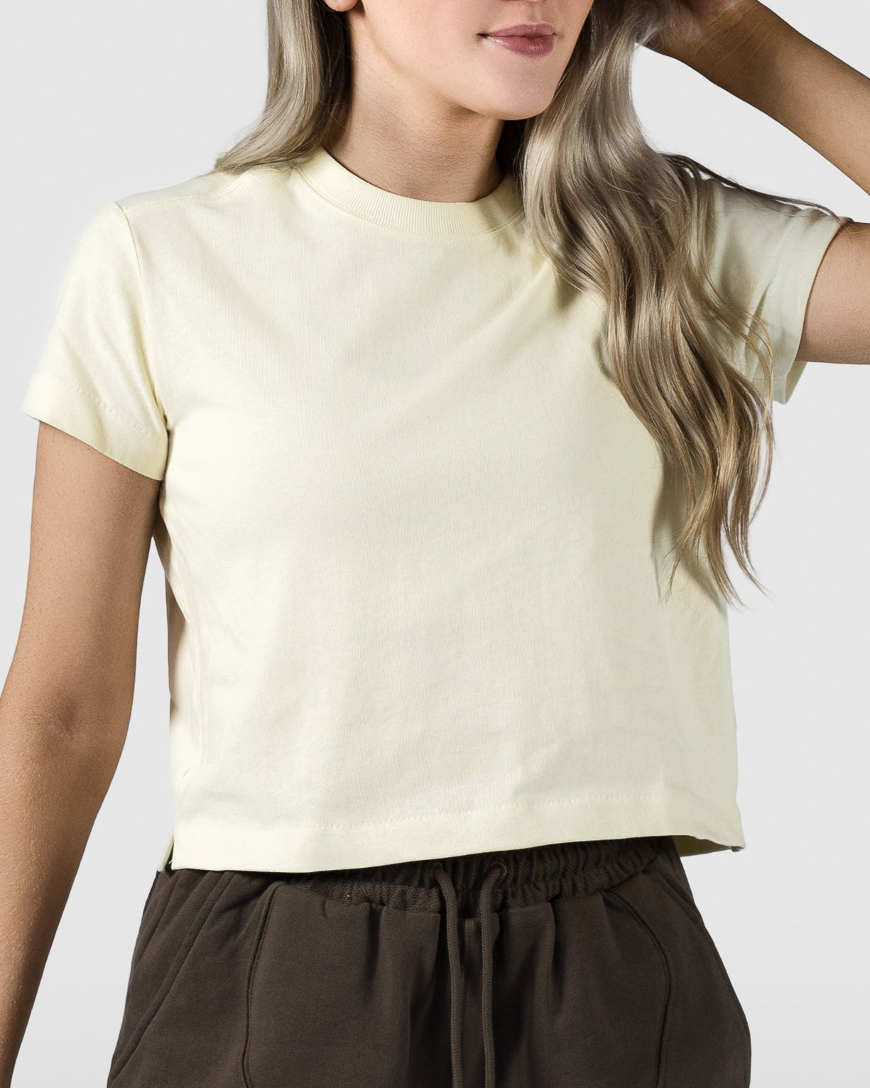 A woman with long, wavy blonde hair wears the BEARDEDGOAT Womens Valley Tee in light yellow and dark brown drawstring shorts, standing against a plain light gray background—an ideal look for relaxed summer days.